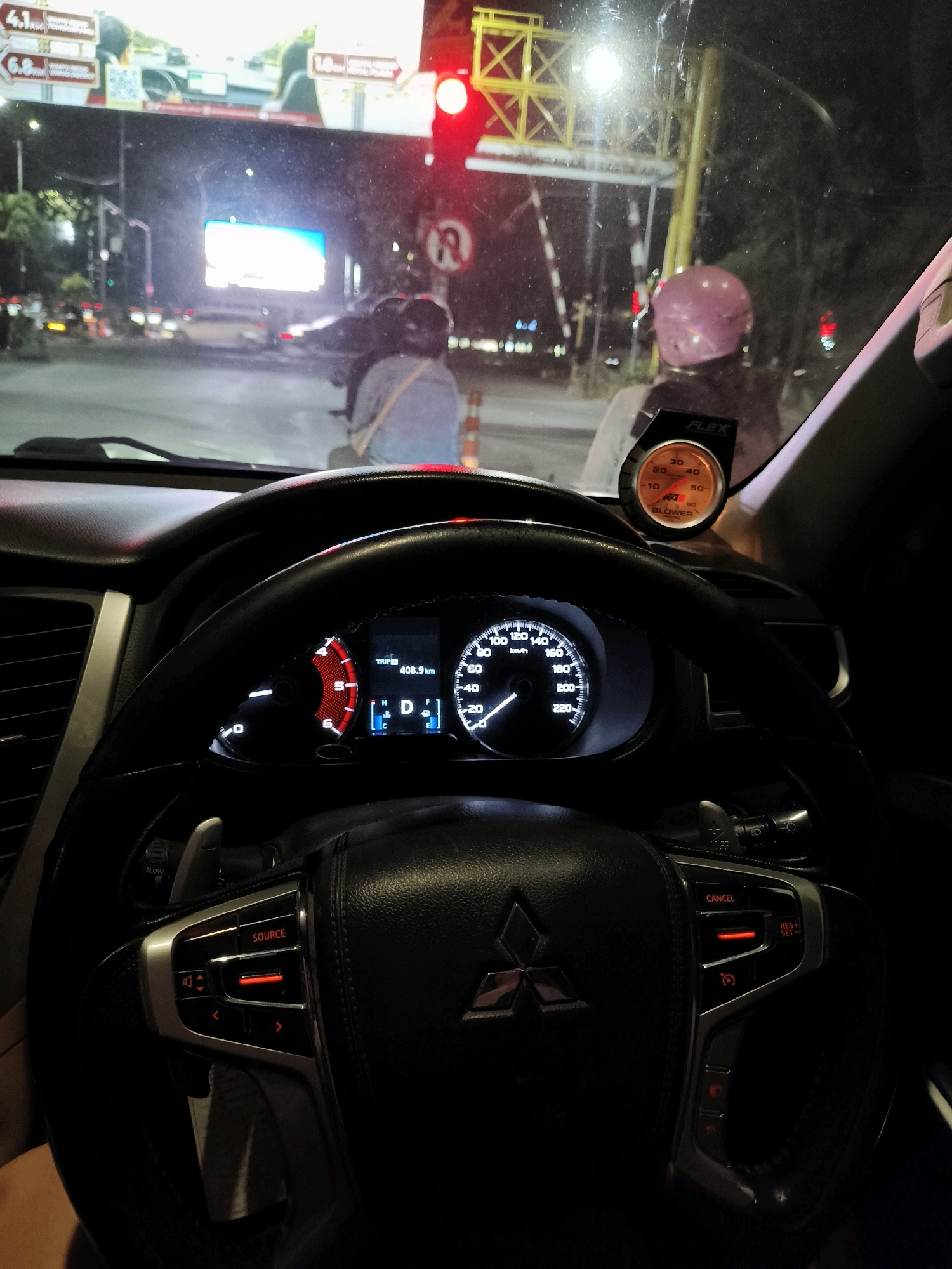 This image captures a view from inside a car in Surabaya, Indonesia, at night. The driver's perspective focuses on the steering wheel and dashboard, with a digital speedometer displaying 40 km/h. To the left of the steering wheel, a partial image of a digital odometer reads "TRIP 408.9 km". A boost gauge, labeled "FLEX" and showing a reading around 30, is mounted on the dashboard.
Outside the car, visible through the windshield, is a traffic scene at an intersection. A red traffic light is illuminated, and a sign with a "no U-turn" symbol is visible. Several vehicles are present, including motorcycles and cars. One person on a motorcycle, wearing a pink helmet, is in the foreground, and another person in a light-colored shirt is further back on a motorcycle. A large illuminated billboard is also visible in the background, displaying advertisements. The scene suggests the car is stopped at a traffic light, possibly waiting to turn.