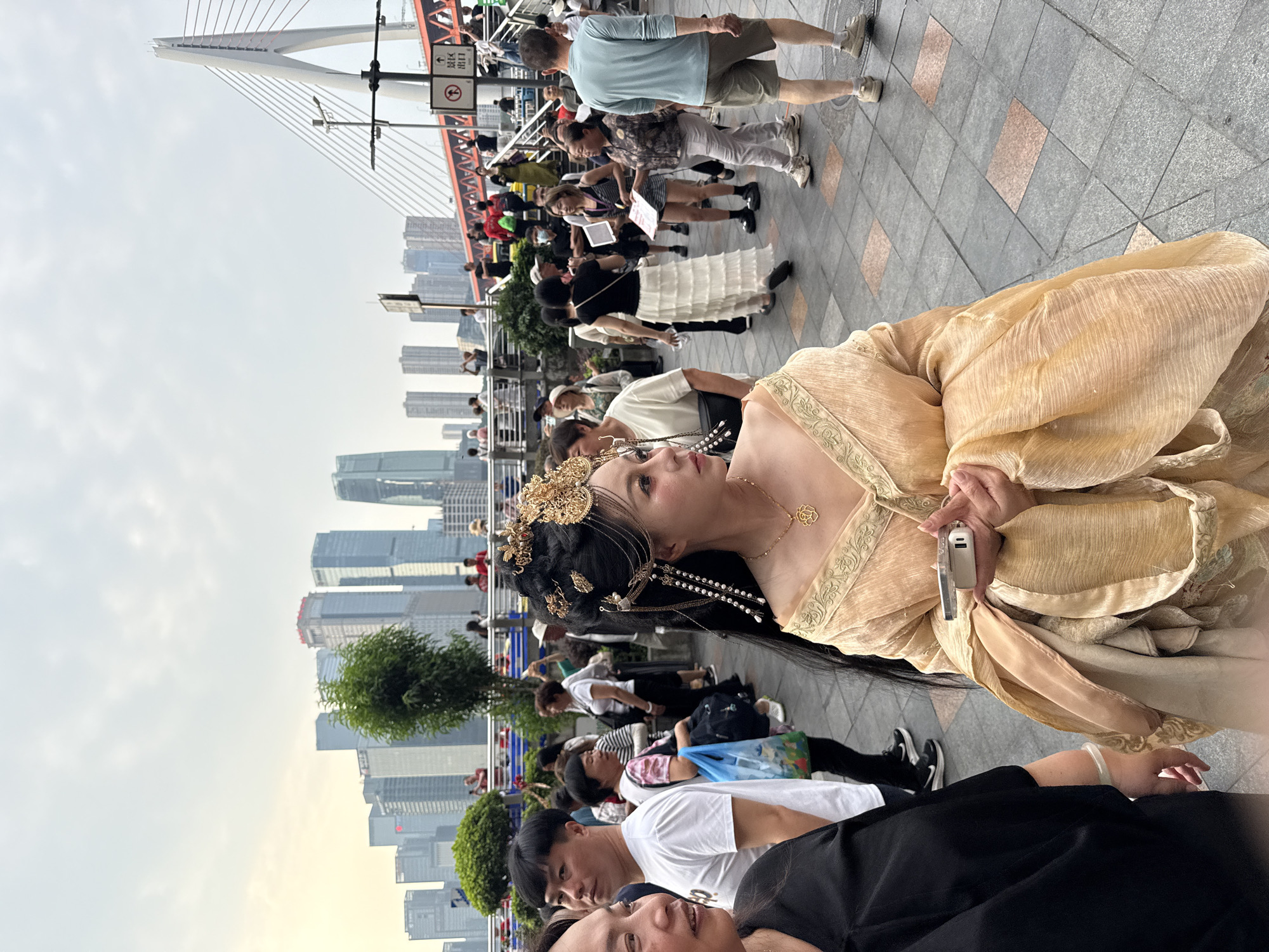 Here's a description of the image:
The image shows a woman in a traditional golden dress with ornate hair decorations holding a small electronic device, possibly a phone or camera, in a public setting. She's positioned in the foreground, slightly angled towards the camera. The background includes a crowd of people, and in the distance, a bridge with prominent cables and modern city buildings, indicating an urban environment. Based on the city skyline and bridge architecture, this image was likely taken in Yuzhong District, Chongqing, China, near the Dongshuimen Bridge. The weather appears overcast, as the sky is cloudy.