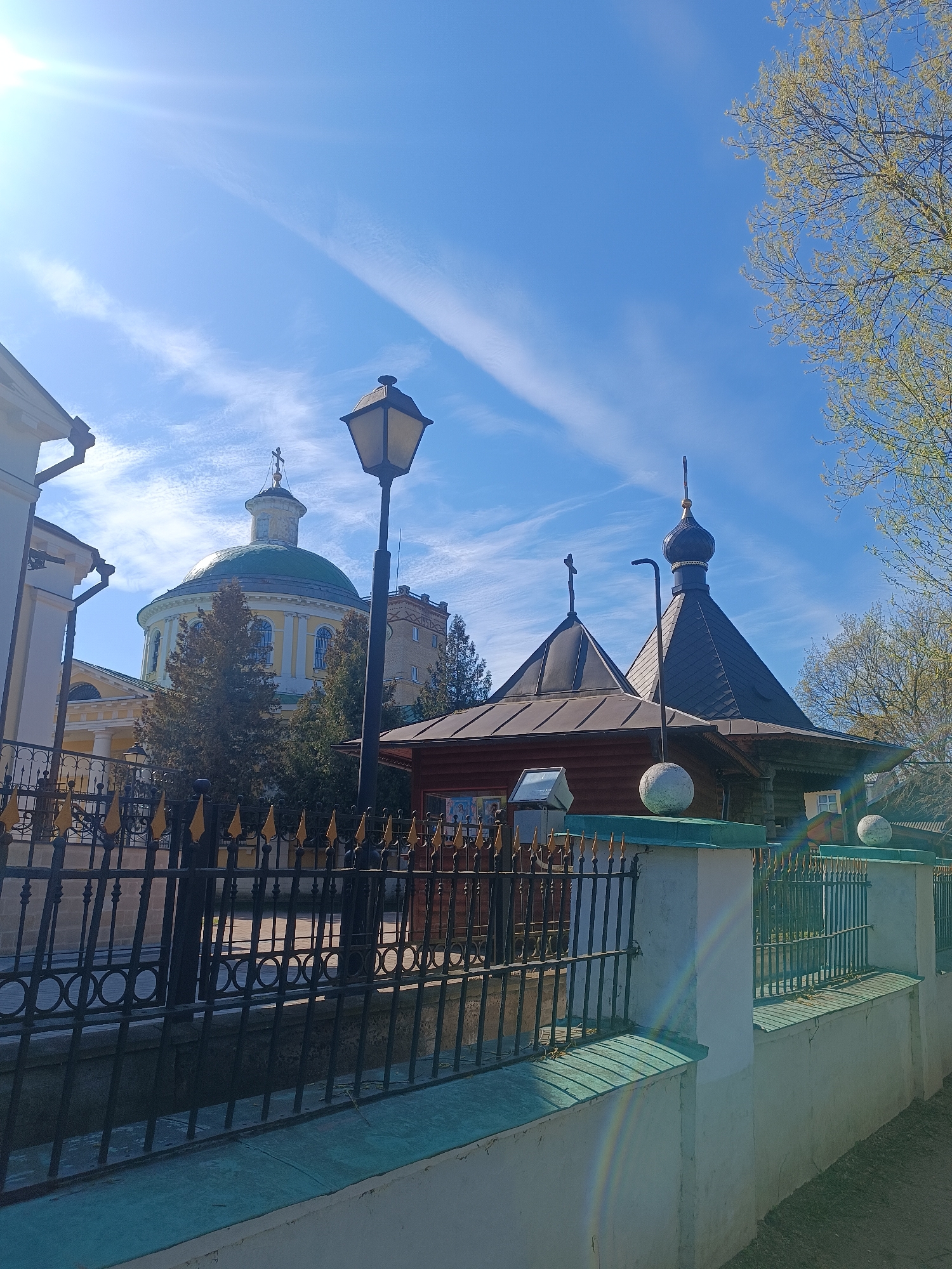The image shows a street-level view of structures within a fence in Moscow, Russia. A large building with a green dome is visible behind a smaller, dark-roofed wooden building. A black metal fence with gold-colored spear-shaped tips runs along the front of the image, atop a white wall with a green-trimmed edge. A lamppost stands near the center of the frame. The sky is blue with light clouds. A section of tree branches is visible on the right side of the image.