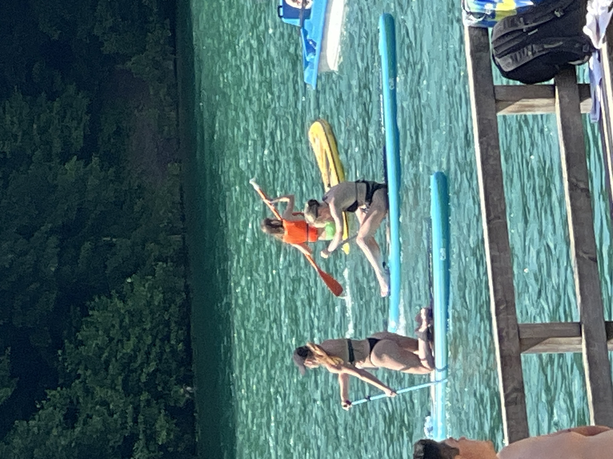 The image depicts a sunny day on a lake, with several people enjoying various water activities. In the foreground, a wooden railing separates the viewer from the water. Behind the railing, three individuals are visible on paddleboards. One person, wearing a black bikini and a black baseball cap, is kneeling on a blue paddleboard and holding a paddle. Another person, wearing a black bikini, is sitting on a blue paddleboard. Further in the background, a third person, wearing an orange life vest and green shorts, is paddling a yellow inflatable. A blue boat with a white hull is also present on the water. The lake is surrounded by lush green trees, suggesting a natural or park-like setting. The water is a vibrant turquoise color, reflecting the bright sunlight. The overall scene conveys a sense of leisure and outdoor recreation. This scene is likely at the Vingo Park's recreational lake in Vilnius, Lithuania, known for its water sports and leisure activities.