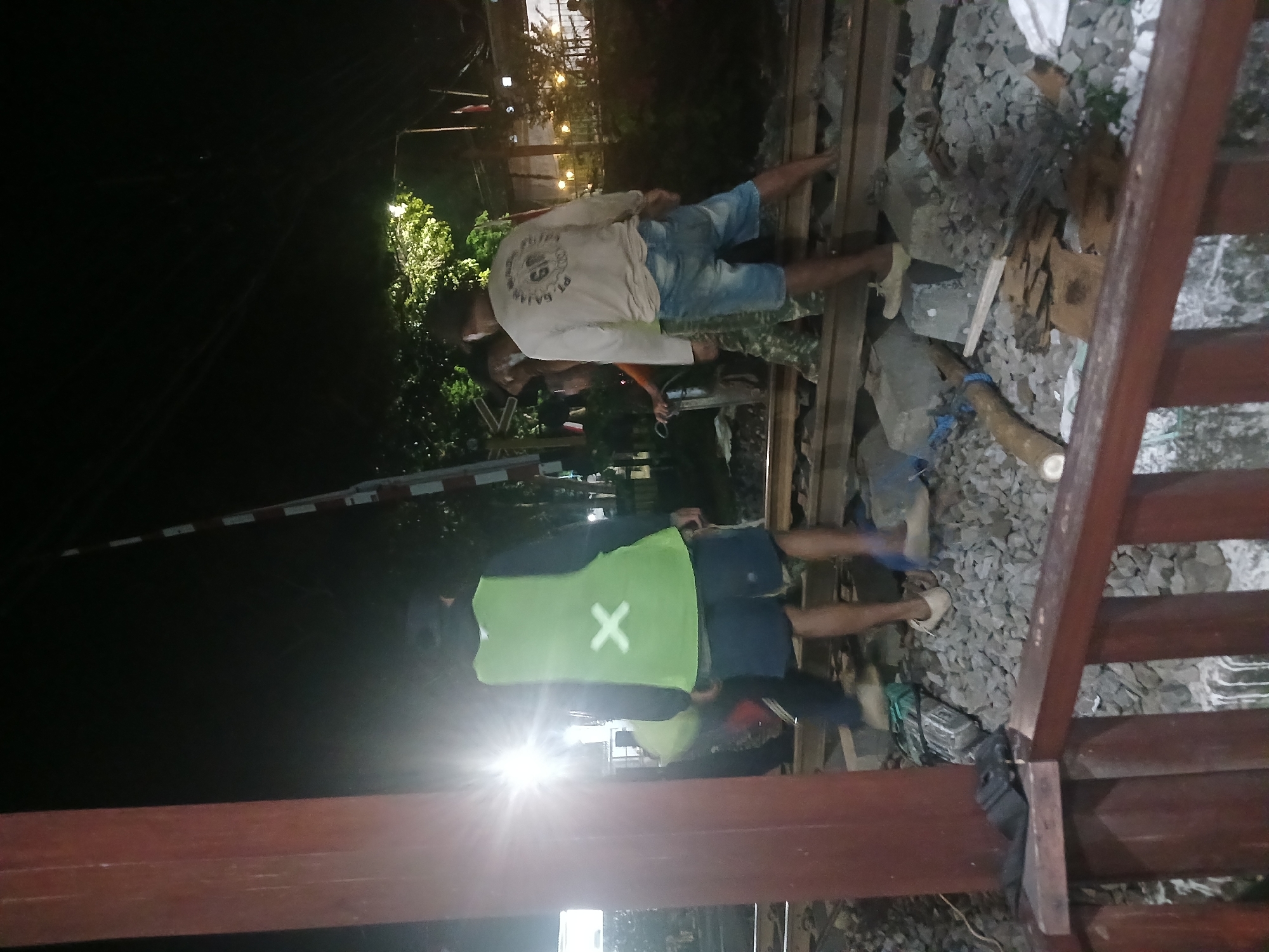 This is a night-time, ground-level, outdoor photograph taken at a railway crossing in Banyuwangi, Indonesia. The scene depicts at least three individuals, likely railway workers, engaged in activity on or around the train tracks.
The individual in the foreground, facing away from the camera, is wearing a bright lime green vest with a white 'X' marking on the back, dark blue shorts, and light-colored shoes. To their left, another person is partially visible, also in work attire. To the right of the first individual, another person is seen from the back, wearing a long-sleeved light-colored shirt with text and graphics, and denim shorts. This person appears to be standing on the railway tracks.
The environment consists of the railway tracks themselves, with gravel ballast. There are wooden support beams and what appears to be a railway crossing barrier pole with red and white markings in the background. Trees and foliage are visible behind the tracks, and some artificial lighting is present, including a bright, intense light source emanating from the left side of the frame, suggesting a powerful work light or flash. The overall lighting conditions indicate it is dark, with the primary illumination coming from artificial sources. The area appears to be some form of railway maintenance or operational site.