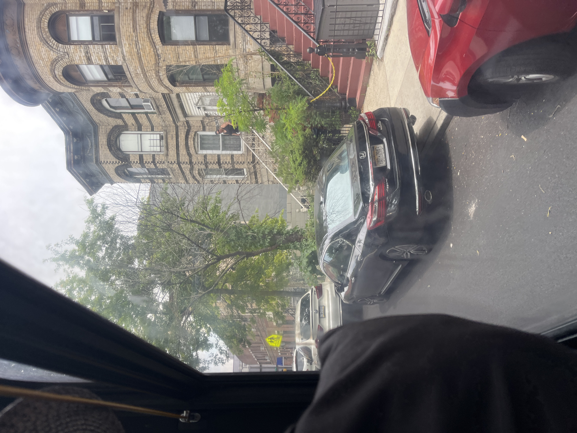 The image shows a street scene in what appears to be a residential area of New York, USA. In the foreground, the interior of a vehicle is visible, with a person's arm and shoulder covered in black fabric. The perspective suggests the photo was taken from inside this vehicle, looking out through the sunroof.
Outside, a row of brick apartment buildings with arched windows lines the street. A red car is parked to the right, and a black car is parked in front of it. A person wearing a dark tank top and shorts is leaning over a white railing on the stairs of one of the buildings. Yellow caution tape is strung across the stairs. Trees and greenery are present along the sidewalk and in front of the buildings.
Further down the street, a yellow school crossing sign is visible, indicating the presence of a school or a school zone. The sky appears overcast, suggesting a cloudy day. The overall impression is a snapshot of everyday life on a New York City street, possibly capturing a minor incident or construction work due to the caution tape.