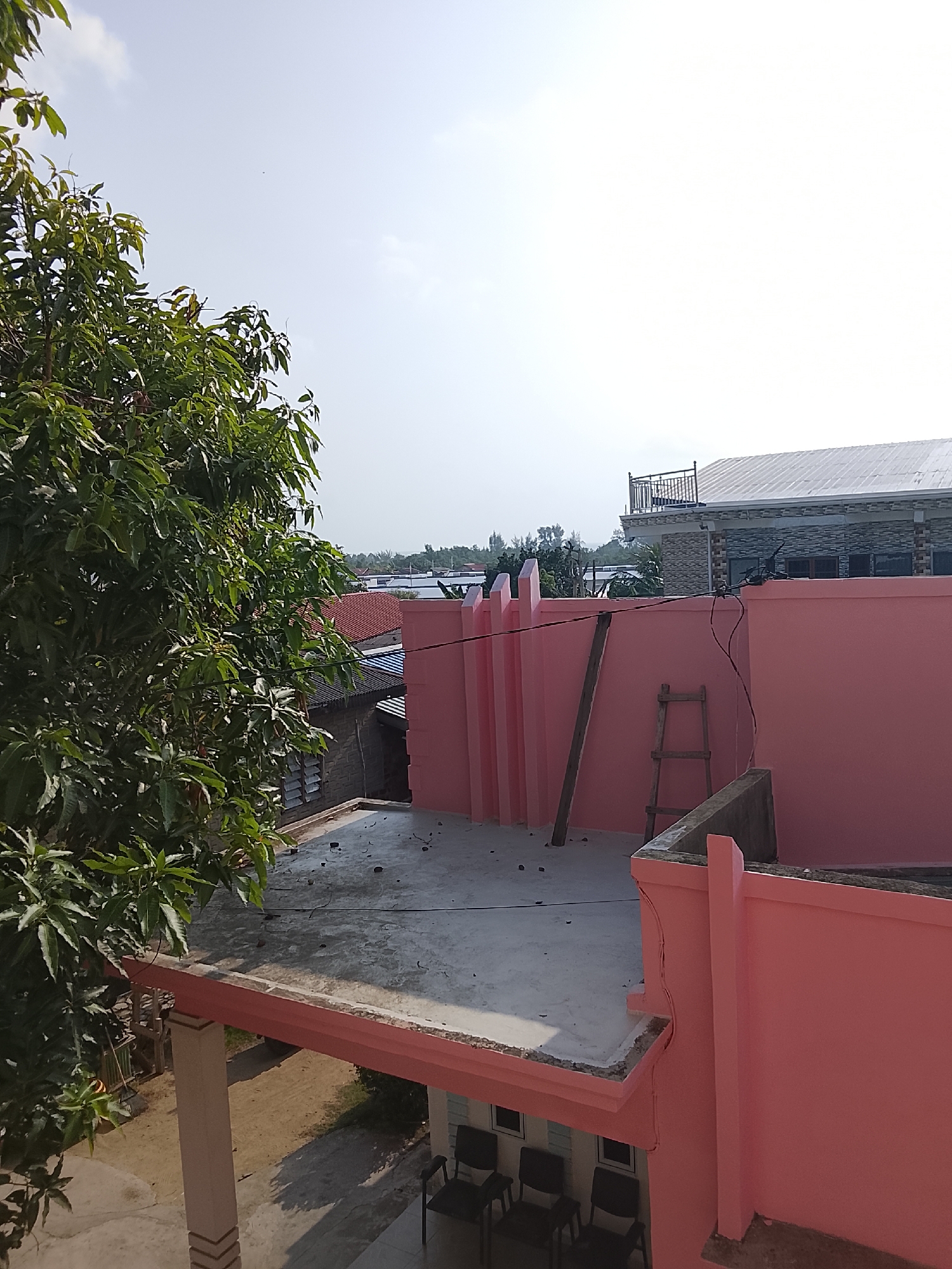 The image captures a residential area in Tanjung Balai Karimun, Indonesia, under a bright, hazy sky. The foreground is dominated by the vibrant pink exterior walls of a building, accented by a concrete roofline. A large, leafy green tree branches into the frame from the left, its foliage obscuring parts of the scene.
On the flat, gray concrete rooftop, a simple wooden stepladder leans against the pink wall. Adjacent to it, a long, narrow piece of wood is propped up. Scattered debris, appearing as small dark specks, is visible on the rooftop. Beyond the pink structure, other buildings are visible, including one with a red-tiled roof and another with a gray, patterned facade topped by a white, corrugated roof. A balcony with a decorative metal railing sits atop this building. The background shows a line of trees and vegetation, suggesting a semi-urban or suburban setting.
The lower part of the image reveals a section of the building's ground level, featuring pillars supporting an overhang. Black chairs are arranged on what appears to be a tiled floor underneath this overhang. The overall impression is one of a sunny day in a residential area undergoing construction or maintenance.
