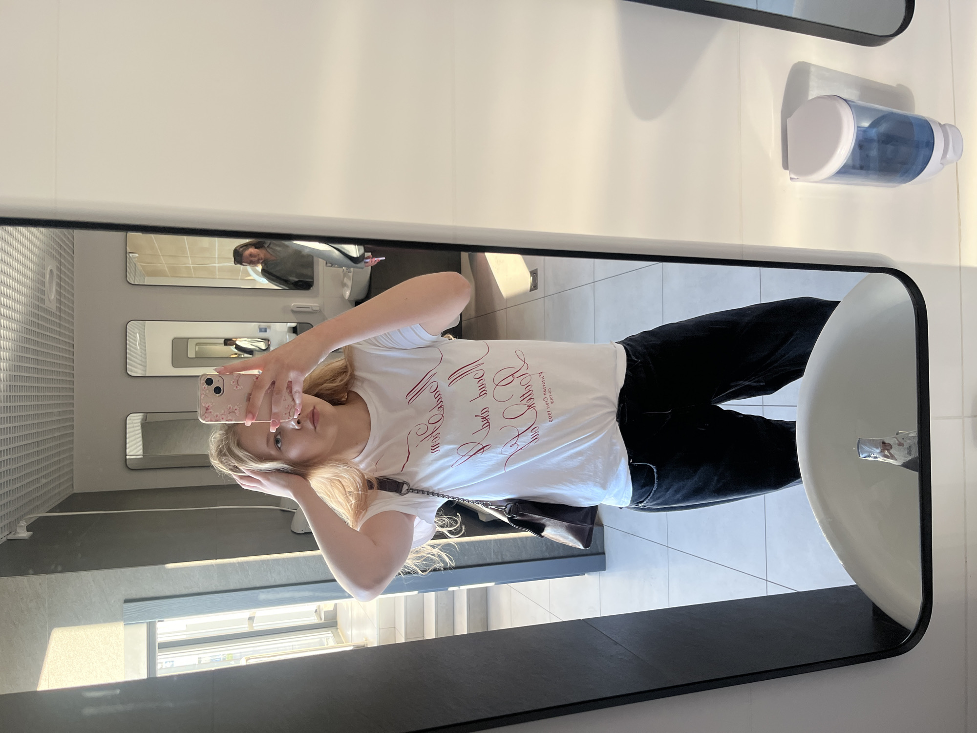 A young woman with blonde hair is taking a mirror selfie in a public restroom. She is wearing a white t-shirt with red script writing and black pants. She has her hands on her head and is looking at the camera. In the reflection of the mirror, another woman is visible in the background. The restroom has tiled walls and floors, several mirrors, and sinks. A soap dispenser is mounted on the wall. The scene appears to be set in a modern, clean public facility. This image was taken in the city of Perm, Russia.