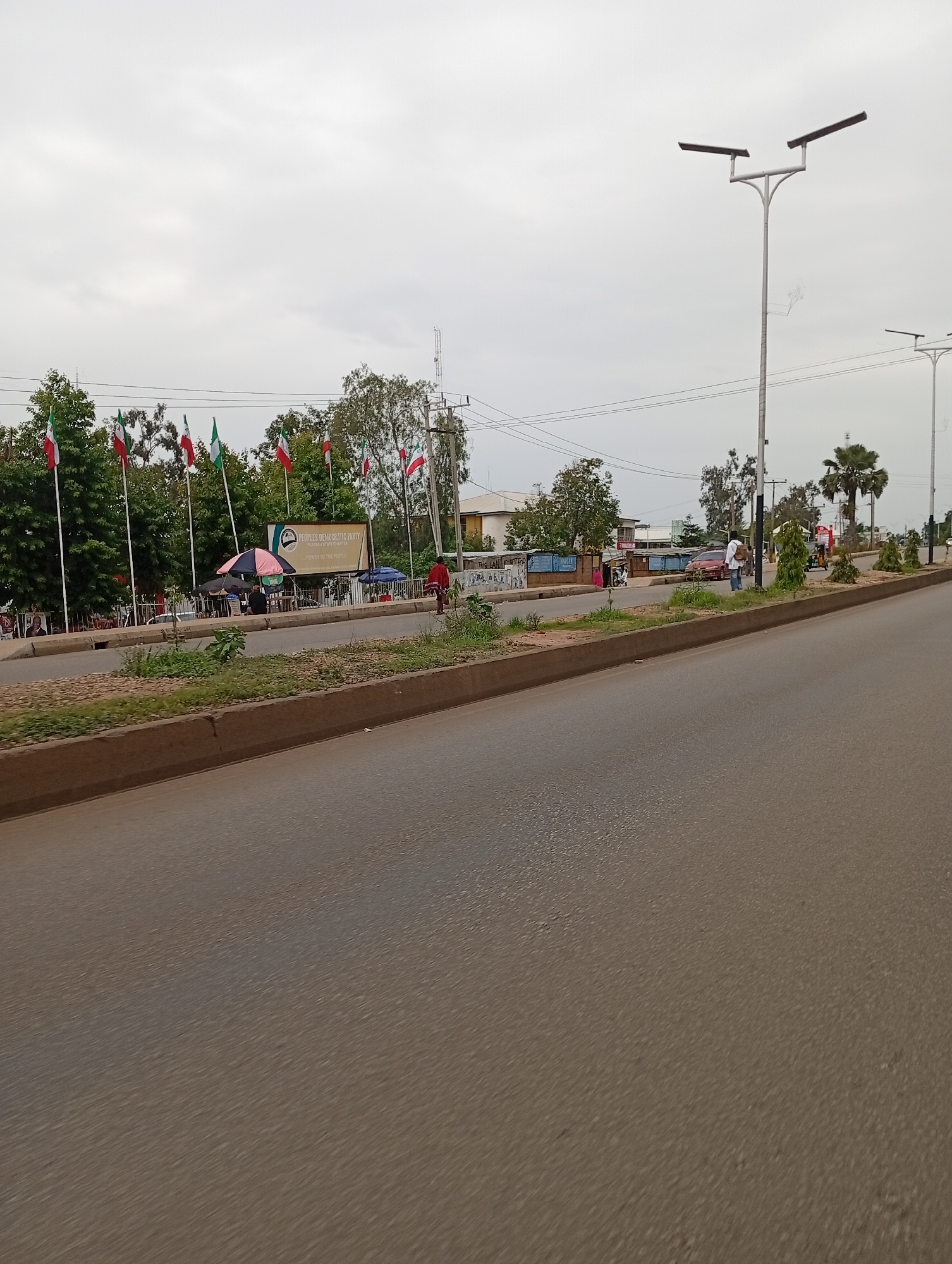 The image shows a street scene in Jos, Nigeria. On the left side of the road, there is a row of flags planted in front of trees and a building. The building has a sign that reads "PEOPLES DEMOCRATIC PARTY PLATEAU STATE CHAPTER". There are several people sitting under umbrellas in front of the building, and some are walking along the sidewalk. On the right side of the road, there are two lampposts, one in the foreground and one in the background. There are also several palm trees and other vegetation. The road itself is paved and appears to be in good condition. The sky is overcast.