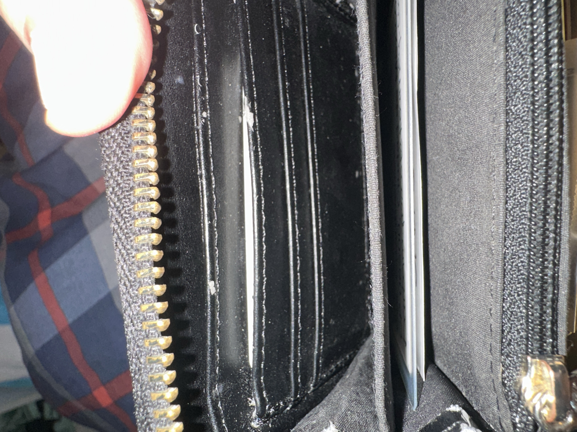 This close-up, slightly overhead image captures the opened interior of a black zippered wallet or purse, held by a person whose thumb is visible in the upper left corner. The wallet features multiple vertical card slots, made of a smooth, dark material, likely synthetic or leather. Several card slots are visible, mostly empty, though the white edge of at least one card is discernible within a slot towards the right.
Small white specks, resembling dust or minor debris, are scattered across the black surfaces of the card slots and along their seams, indicating typical wear and usage. A prominent metal zipper with gold-colored teeth and a dark fabric tape runs along the left edge of the wallet. Further inside, to the right, another internal zippered compartment is visible, with a metallic zipper pull partially showing. The internal lining of the wallet appears to be a dark, fine-textured fabric.
In the blurred background, a pattern of red and blue plaid, consistent with a shirt, can be seen. The lighting is bright, creating a significant highlight on the person's thumb. The activity depicted is the holding and opening of the wallet, suggesting an interaction with its contents or an examination of the item. There are no explicit location cues, time of day, weather, or discernible emotions. No visible text is present in the image.