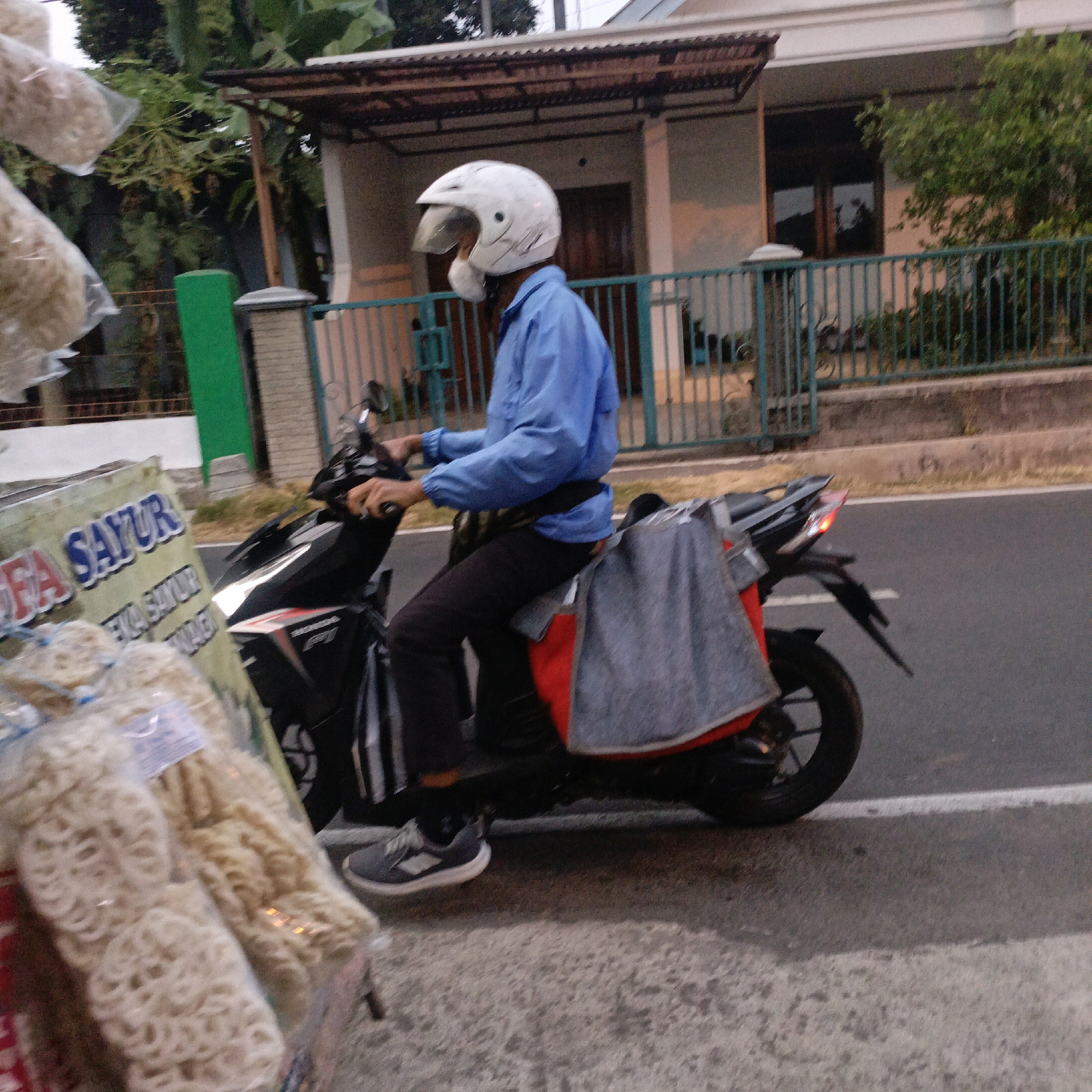 A person wearing a blue shirt and black pants, with a white helmet, rides a black motorcycle. The motorcycle has a red and black decal on its side, and a gray fabric is draped over the rear seat. The person is on a road in front of a house with a brown tiled roof and a teal fence. Bags of snacks are hanging from the left side of the image, and a green pole is visible behind the snacks. The scene is likely in Salatiga, Indonesia, based on the signage.