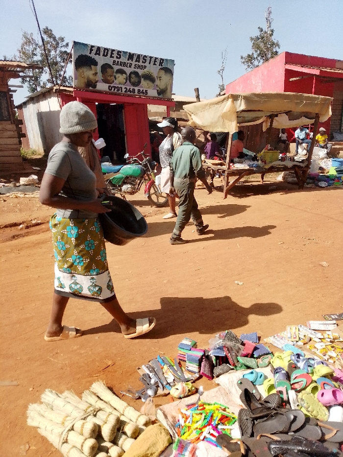 The scene takes place in Lwandeti, Kenya, at coordinates 0.563 latitude and 34.806 longitude. A woman in a grey shirt and a patterned skirt is walking down a dirt road. She is carrying a black plastic basin. In front of her are various items for sale including sandals, brooms, and kitchen utensils. There are also various stalls and shops, including a barber shop called "Fades Master". The barber shop is red and has a sign with a phone number. There is a motorcycle parked in front of the shop. There is another woman walking towards the right of the image, along with two other men. It appears to be a sunny day.