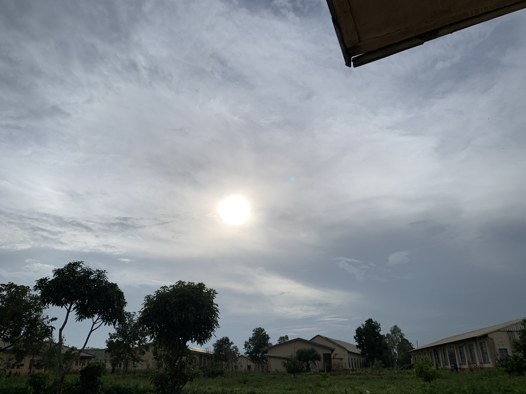 The image captures an outdoor scene in Kuru Karama, Nigeria. The sky is overcast with scattered clouds and the sun is visible as a bright, diffused orb in the upper center of the frame, casting a soft light. In the foreground, there are several trees with dense foliage, some appearing to be mango trees. Behind the trees, a series of light-colored, single-story buildings with sloped roofs are visible. These structures have numerous windows. The ground is covered in lush green grass and some smaller plants. A section of a wooden structure is visible in the upper right corner, appearing to be a roof overhang. The overall impression is one of a rural or semi-rural setting, possibly a school or an institutional compound.