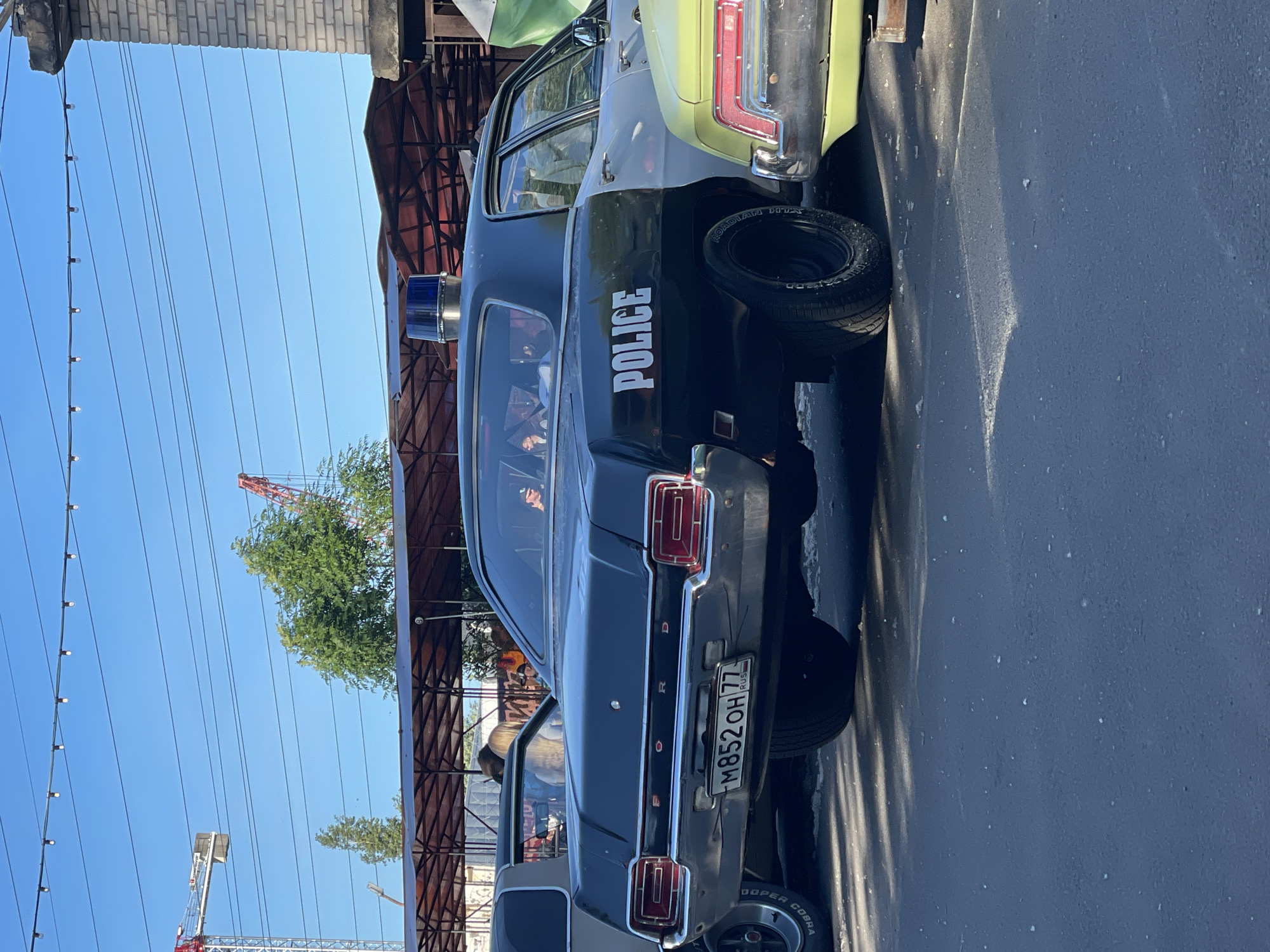 The image captures a vintage black and white police car parked outdoors. The car features a blue light bar on its roof and "POLICE" written in white on its side. The license plate reads "M852OH 77 RUS". In the background, there is a building with brickwork and a canopy structure. A bright blue sky is visible above, with some power lines and a red crane in the distance. The ground appears to be asphalt. The overall scene suggests an outdoor event or exhibition, possibly in Saint Petersburg, Russia, given the license plate format.