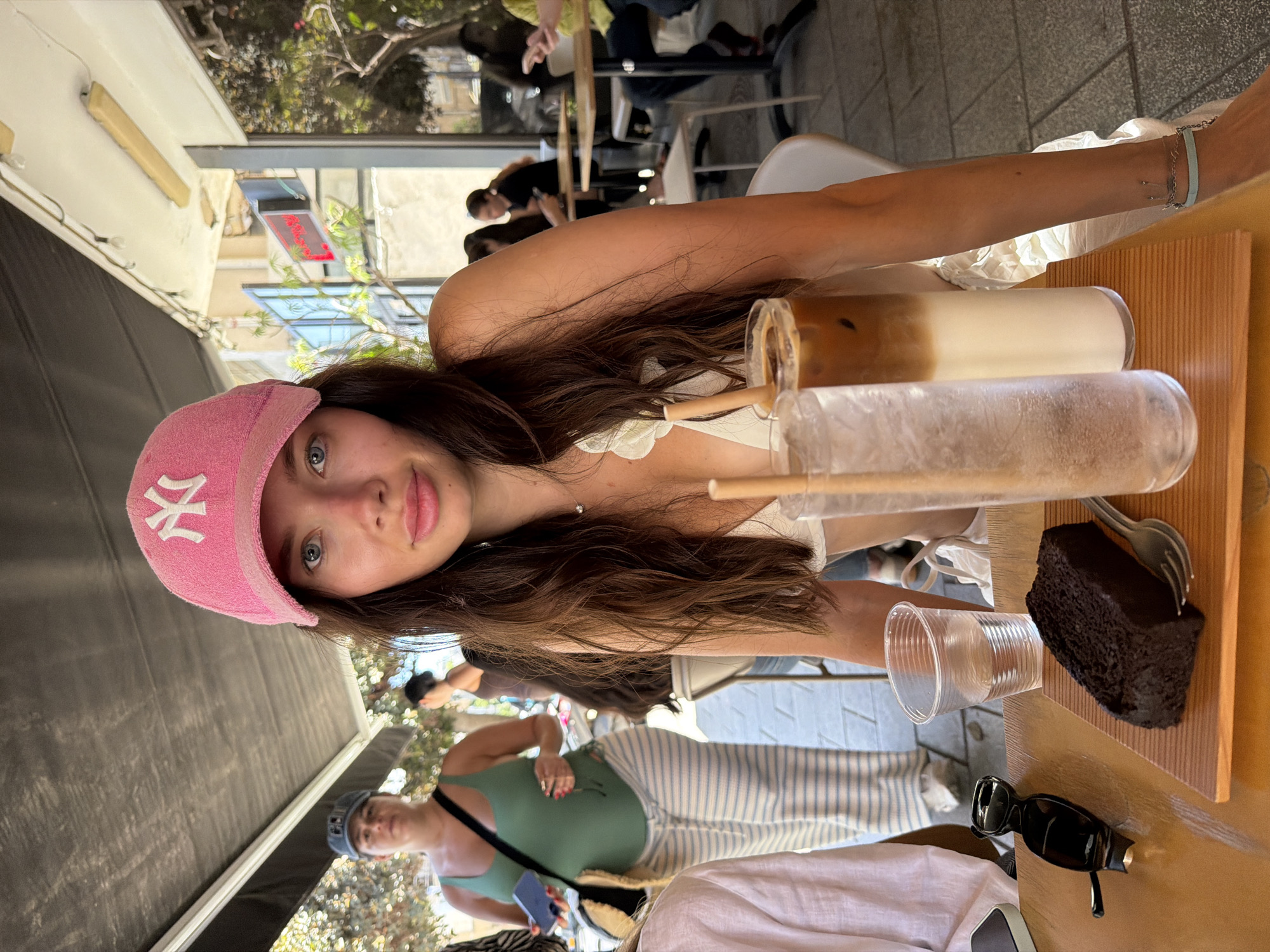 This image captures an outdoor cafe scene in Tel-Aviv, Israel, under a dark awning during daylight hours. In the foreground, a young woman with long, dark brown hair and blue eyes looks directly at the camera. She wears a faded pink New York Yankees baseball cap and a white top with a delicate floral detail on the left strap, revealing a small pendant necklace. On the light wooden table in front of her are several items: a dark chocolate loaf cake resting on a wooden board with a metal fork, a tall glass containing a clear liquid with ice, and another tall glass holding a layered iced coffee drink. A clear plastic cup with water and a pair of black sunglasses are also on the table to the left.
In the mid-ground, slightly to the left, a woman wearing a dark cap, a green tank top, and striped light-colored pants walks across the scene. She holds a dark blue phone in her right hand and carries a striped bag over her left shoulder. Further in the background, other patrons are visible seated at various outdoor tables, some appearing to be engaged with their mobile phones. The background features urban elements including a building with windows, a partially visible red neon sign, and some scattered foliage. The ground is paved, and light fixtures are visible along the underside of the awning.