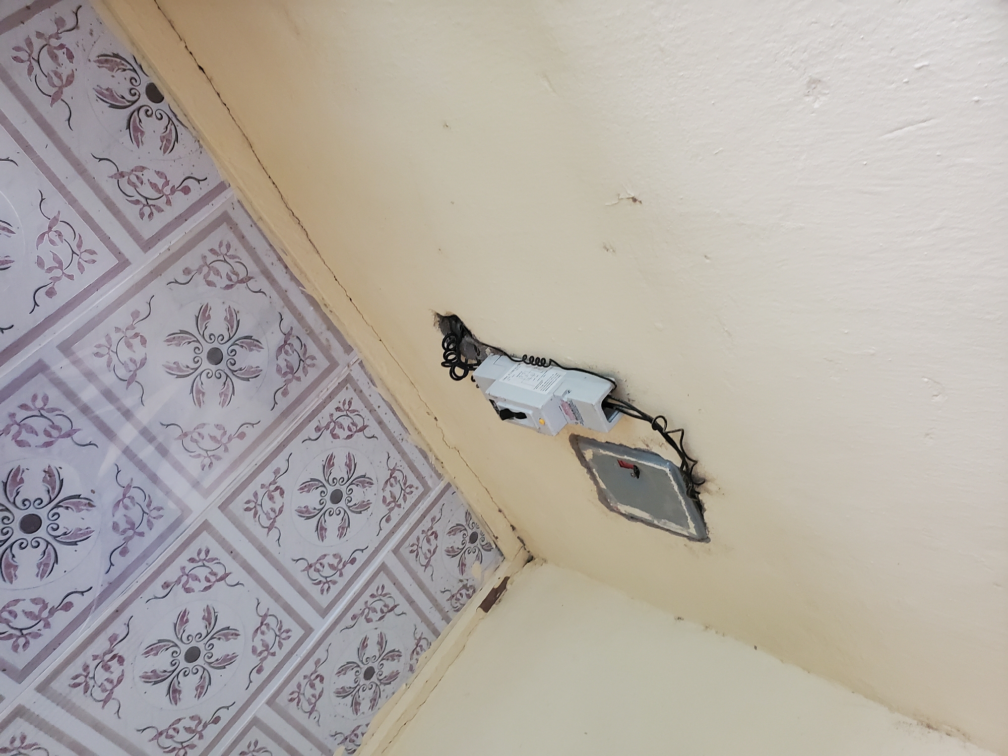 The image shows an interior corner of a room with a textured ceiling and light yellow walls. The ceiling has a tiled pattern with a decorative floral design in shades of pink and purple. The walls appear to be plastered and painted.
Mounted on the wall is an electrical meter and a small distribution box. The meter is white with a display screen and some labels. It is connected by black wiring that enters a rough opening in the wall. The distribution box is rectangular with a grey cover and a red handle or switch visible. The opening around the distribution box and meter appears to be somewhat crude. There are some minor cracks and imperfections visible in the wall and at the junction of the wall and ceiling.
The scene is a typical interior residential or commercial space, possibly a utility area or a room where an electricity meter is located. Given the context of the location provided, this scene is in the city of Jalingo, Nigeria. There are no people visible in the image.