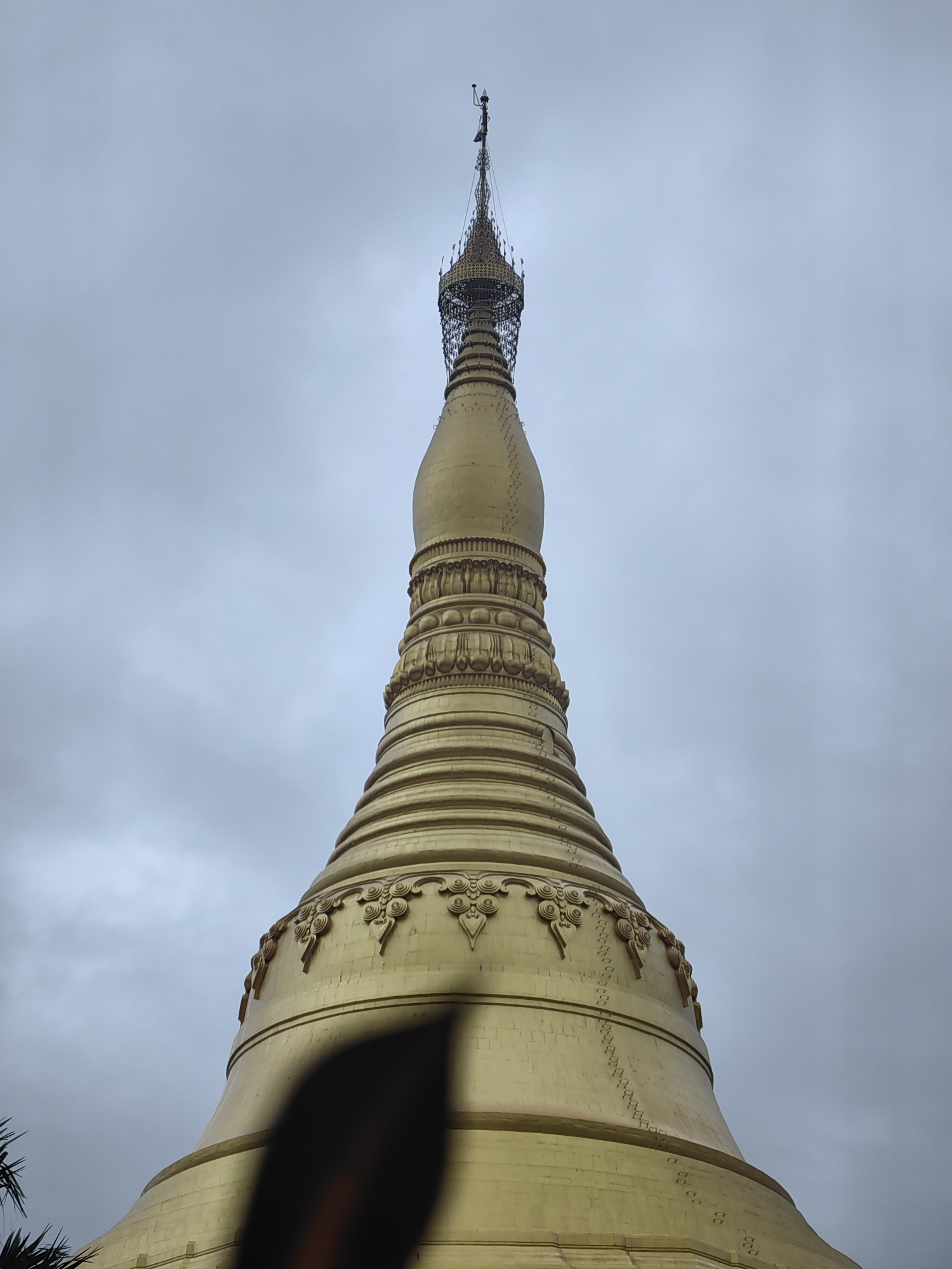 The image shows the upper portion of a golden pagoda against a cloudy sky. The pagoda has intricate detailing on its tiered structure, with decorative bands and circular motifs. A dark, blurry object is in the foreground, partially obscuring the lower part of the pagoda. To the far left, a bit of greenery, possibly palm fronds, is visible. The overall lighting suggests an overcast day. The location is identified as a temple in Mumbai, India.