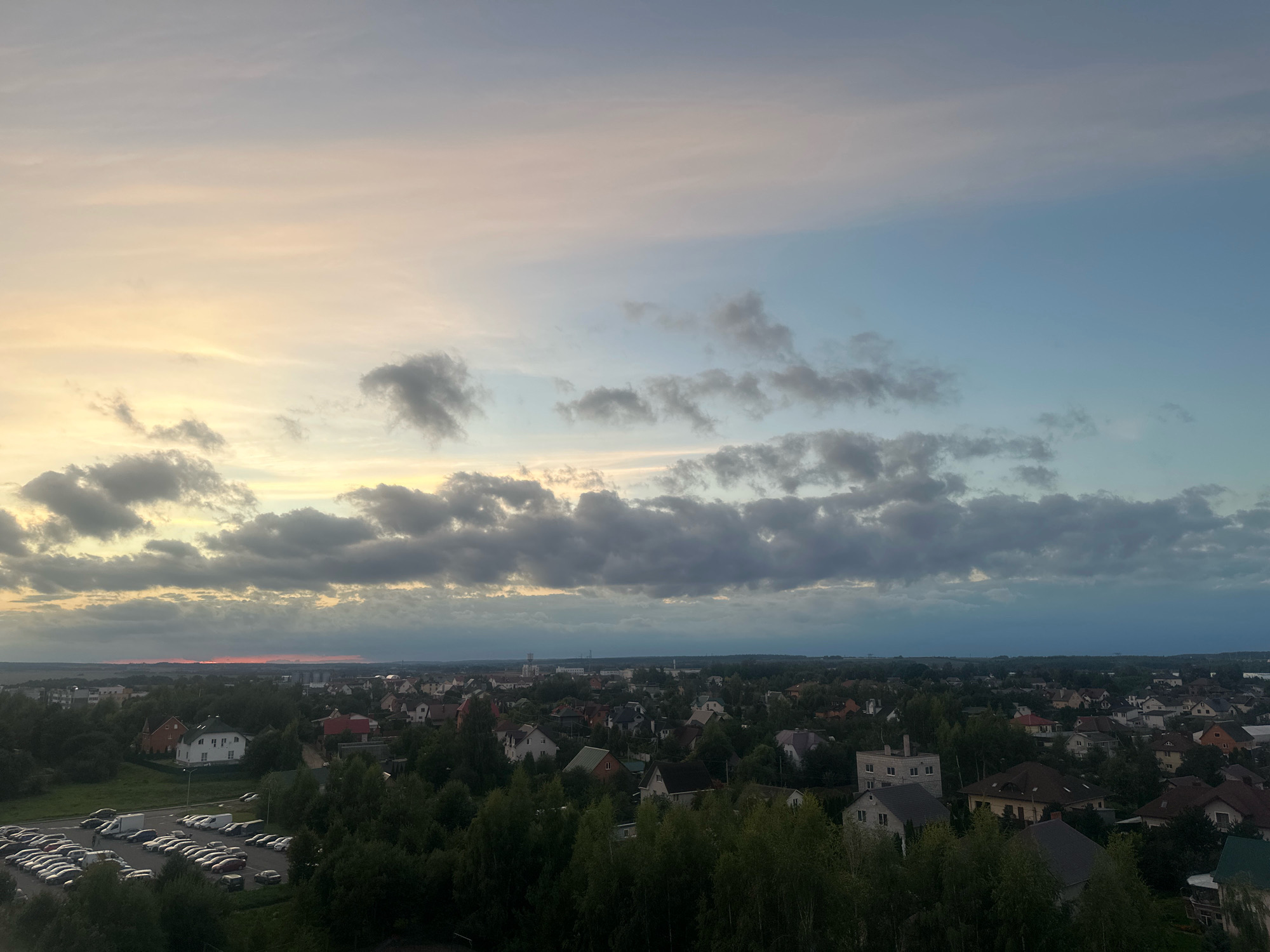 This is an aerial photograph of a suburban area, likely in or near the Шчомысліцкі сельскі Савет in Belarus, taken during the late afternoon or early evening. The sky is a gradient of pastel colors, transitioning from soft yellow and pink near the horizon on the left to a pale blue on the right, with scattered clouds of varying densities. Below the sky, a sprawling residential area is visible, characterized by numerous houses with pitched roofs, many surrounded by lush green trees. A large parking lot filled with many light-colored cars is prominently featured in the lower left quadrant of the image. In the distance, the landscape appears to be a mix of further housing and undeveloped land, with a faint reddish glow on the horizon suggesting the setting sun is obscured by clouds. The overall impression is one of a peaceful, residential evening scene.