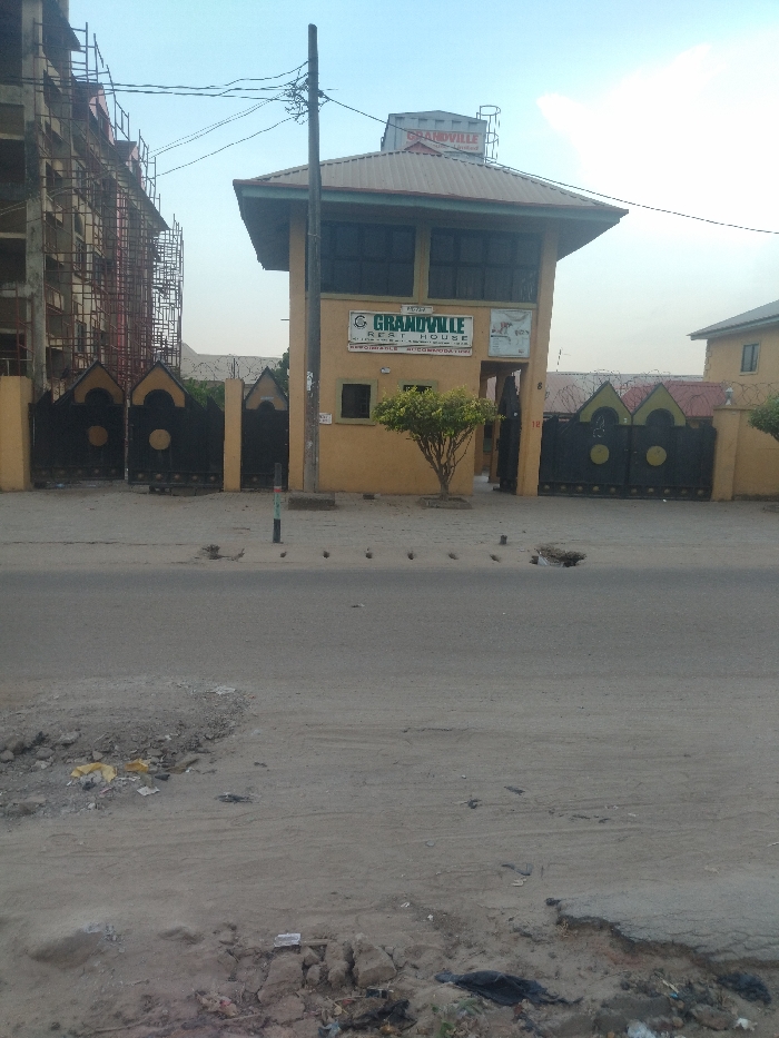 The image shows a street in New Karu, Nigeria. The street is paved with asphalt, but there are several potholes and cracks in the surface. There are several pieces of trash and debris scattered along the edges of the road. In the foreground, there is a small tree. The scene is captured from a street level perspective. There is a building in the background. The building is a tan color and appears to be a hotel or guest house. The building has a sign that reads "Grandville Rest House," "Reasonable Accommodation." The sign is located above a gate with a sign that reads "Entry." There is also a water tank on the roof. The building has several windows and a large, open-air space on the second floor. The building has several windows and a large, open-air space on the second floor. There is a chain-link fence in front of the building. There is a small tree in front of the building. The scene is taken on a cloudy day with a bright blue sky visible. The image was taken from a street level perspective. There are no people in the image. The only subject is the Grandville Rest House building. The image suggests that the building is located on a street with moderate traffic.
