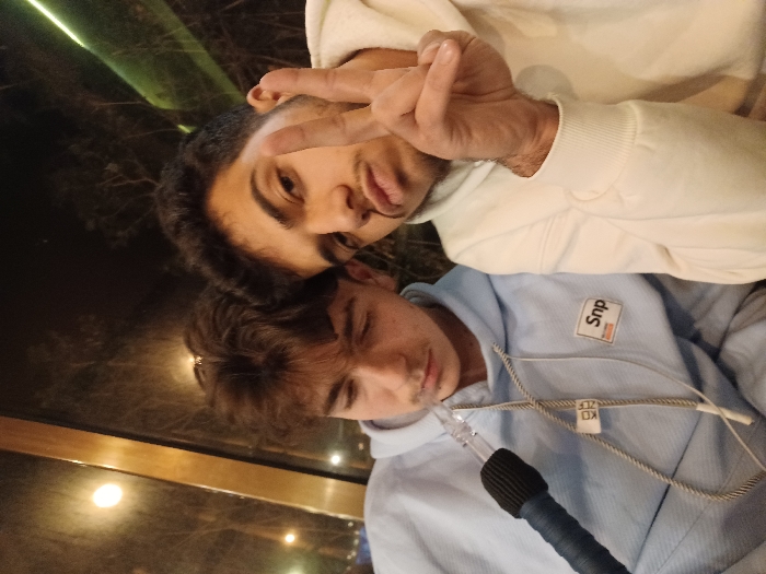 Two young men pose for a selfie. The top man, wearing a white hoodie, is making a peace sign with his fingers. The other man, below, is wearing a light blue hoodie with a "Snp" patch on the chest and appears to be holding a hookah. The background is a dimly lit, blurry scene, possibly outdoors with greenery and a window. The image is slightly rotated.