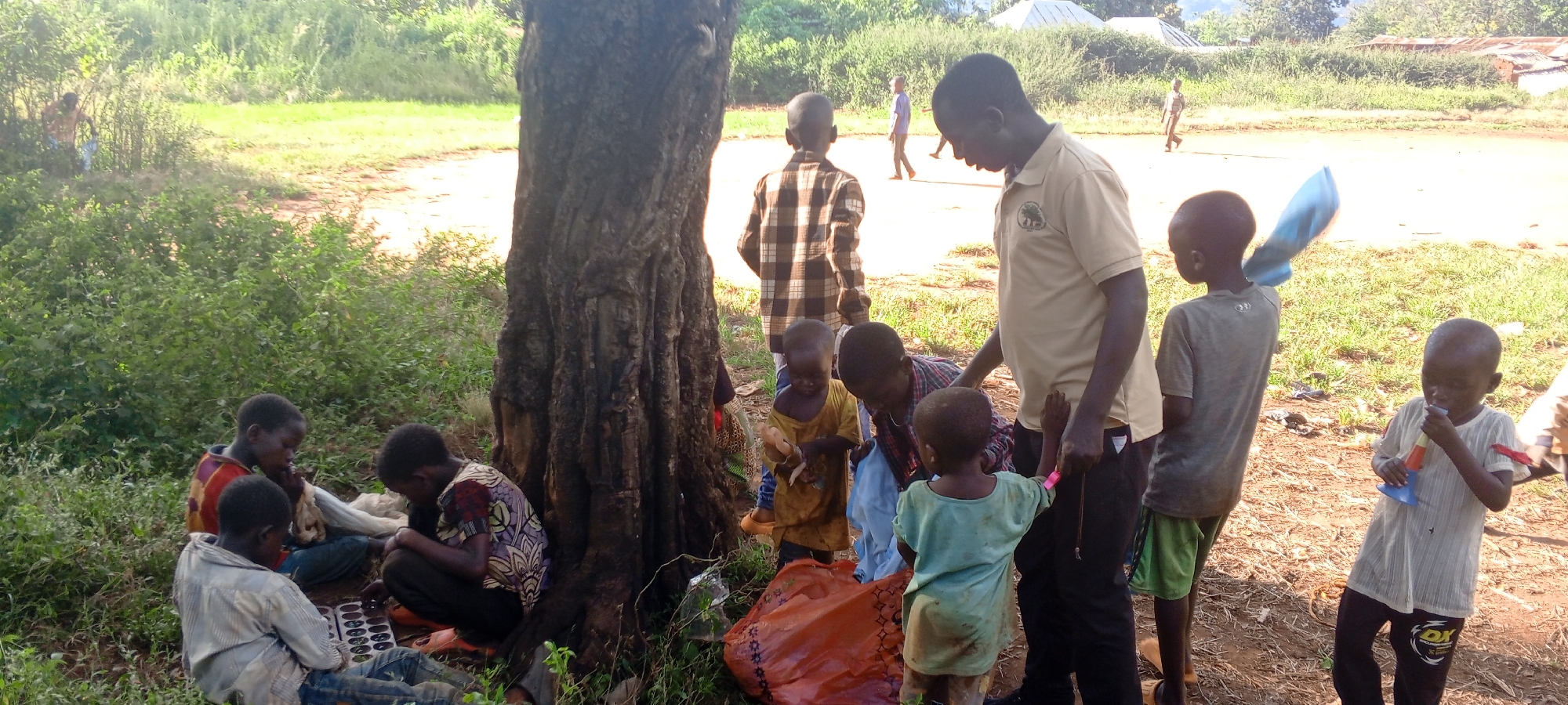 In Ngalula, Democratic Republic of the Congo, a group of children gather near a large tree in a grassy area. Some children sit on the ground, playing a board game on a tray with small depressions or holes. Others stand near an adult male who wears a beige shirt with a logo; he appears to be interacting with the children. A large orange bag lies on the ground near the tree. In the background, other figures are visible walking on what appears to be a dirt road or path. Some buildings are seen further in the background. Vegetation includes bushes and trees.