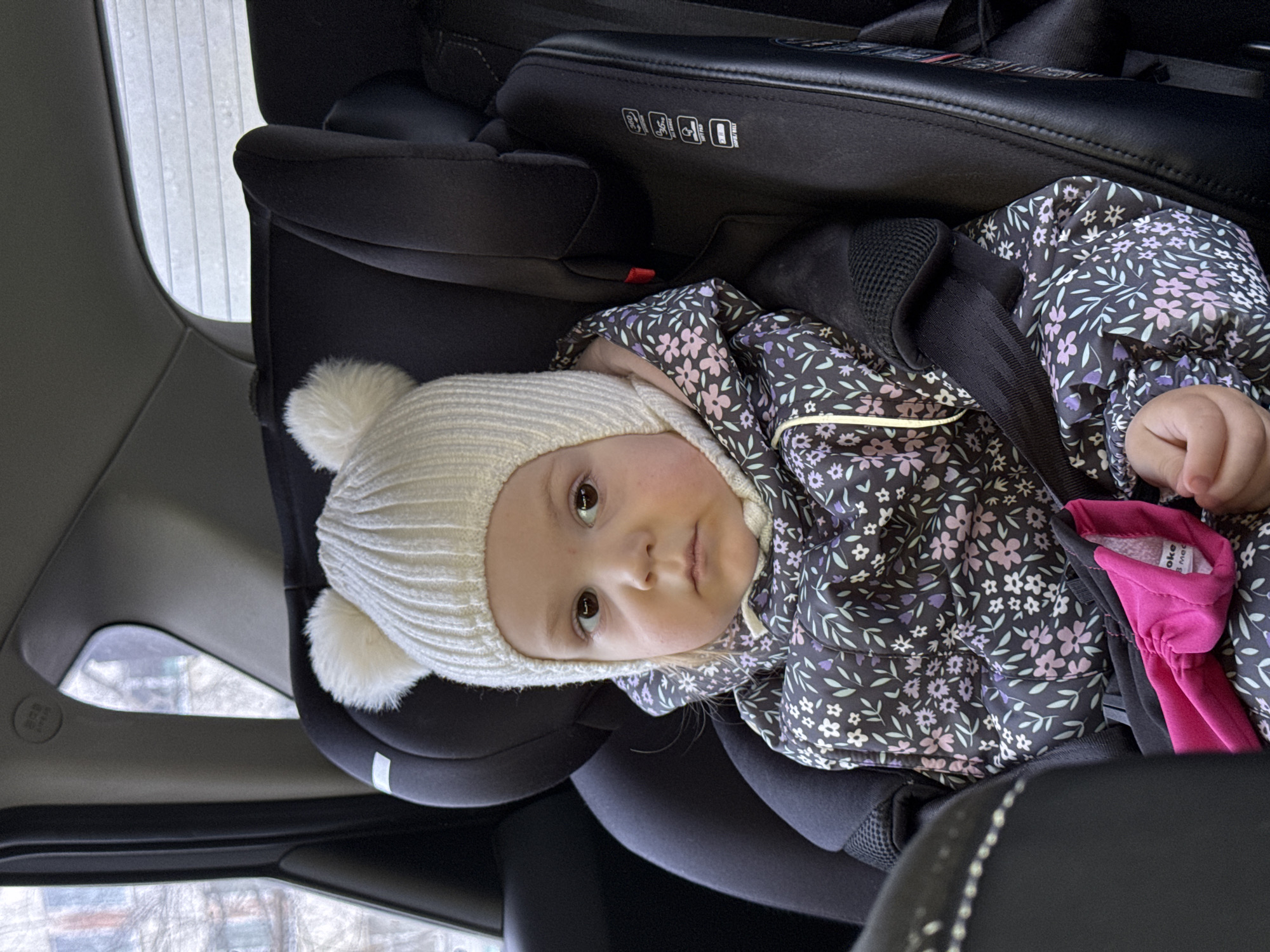 A young child, approximately a toddler, is depicted seated in a black car safety seat within a vehicle. The child is wearing a white knit beanie topped with two white pom-poms and a dark-colored winter suit featuring a pattern of pink and purple flowers and green leaves. The child has light-colored hair, brown eyes, and looks directly forward with a neutral expression. A bright pink fabric item, possibly a mitten, is visible on the child's lap. The car seat has visible safety harnesses and text labels indicating features like "360 rotation" and "ISOFIX." Through the left side window of the vehicle, a building with vertical window panes and faint outlines of tree branches are visible, indicating an urban or suburban environment. The rear window shows a blurry, light-toned background with horizontal elements. The specific location within the city of Chaikovsky, Russia, cannot be determined from the visual information provided.