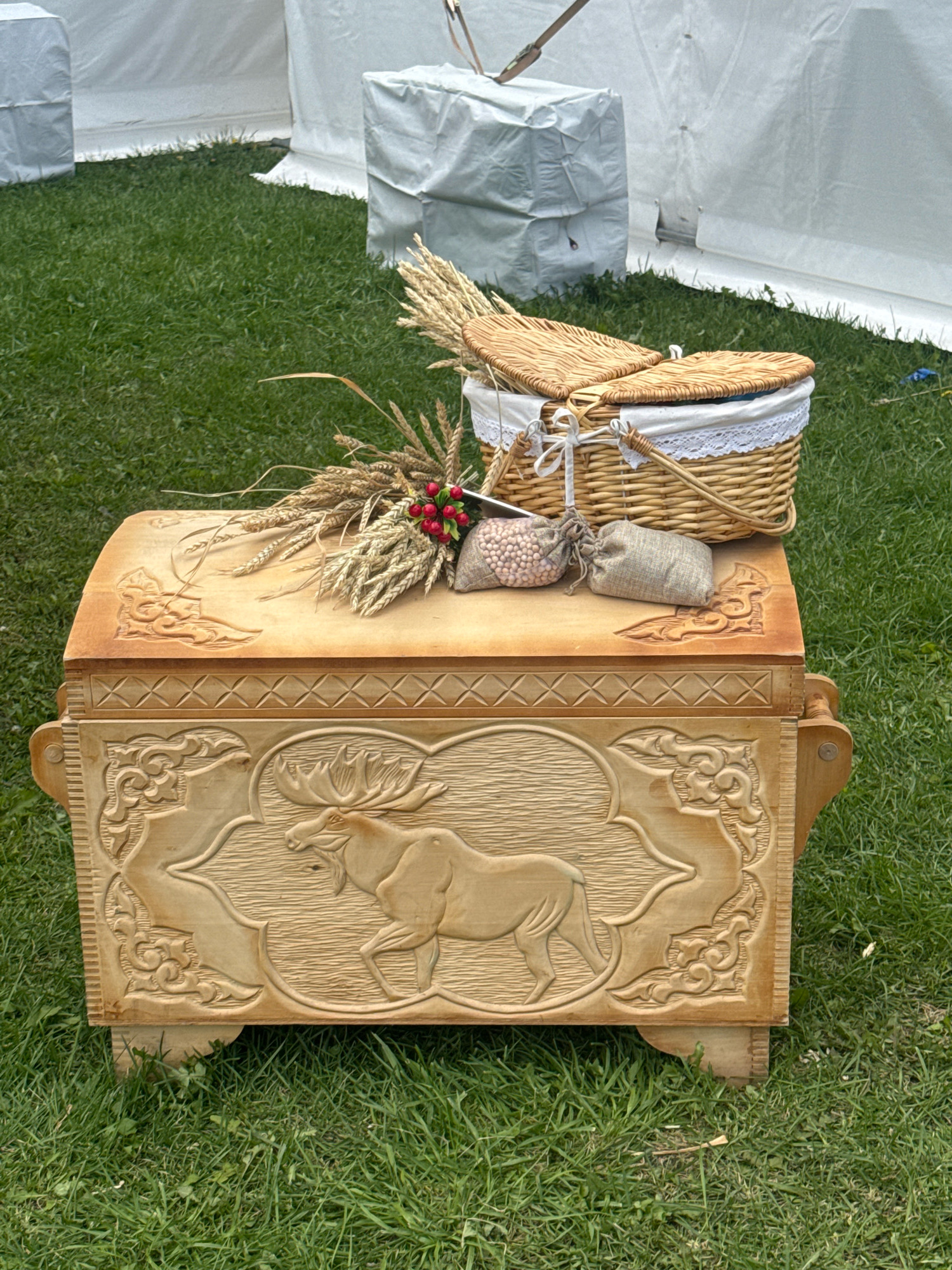 The image shows a wooden chest with carved reliefs of an elk and ornamental patterns. On top of the chest are a wicker basket, a bundle of wheat stalks, and two small burlap sacks. The chest and its contents are placed on a grassy area, with white tent-like structures in the background. The scene appears to be an outdoor setting, possibly a fair or market.