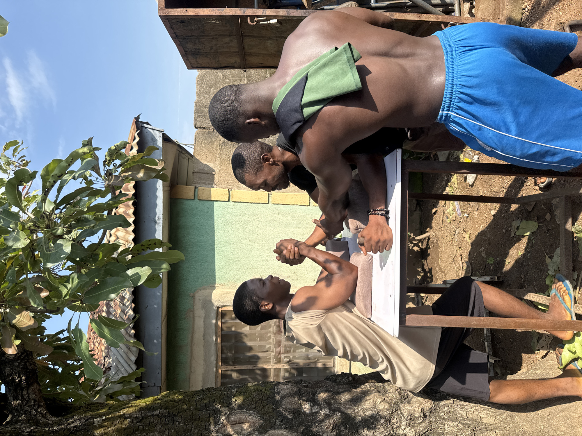 Three Black men are engaged in an arm-wrestling match outdoors. Two men are positioned opposite each other at a table, their arms clasped, in the midst of their competition. The third man stands behind one of the competitors, observing the match. The scene is set in Akwanga, Nigeria, indicated by the architectural style of the buildings and the surrounding vegetation. The background features a green-painted building with a corrugated metal roof and a large tree on the left, casting shadows on the ground. The ground is dirt, scattered with fallen leaves. The sky is clear and blue.