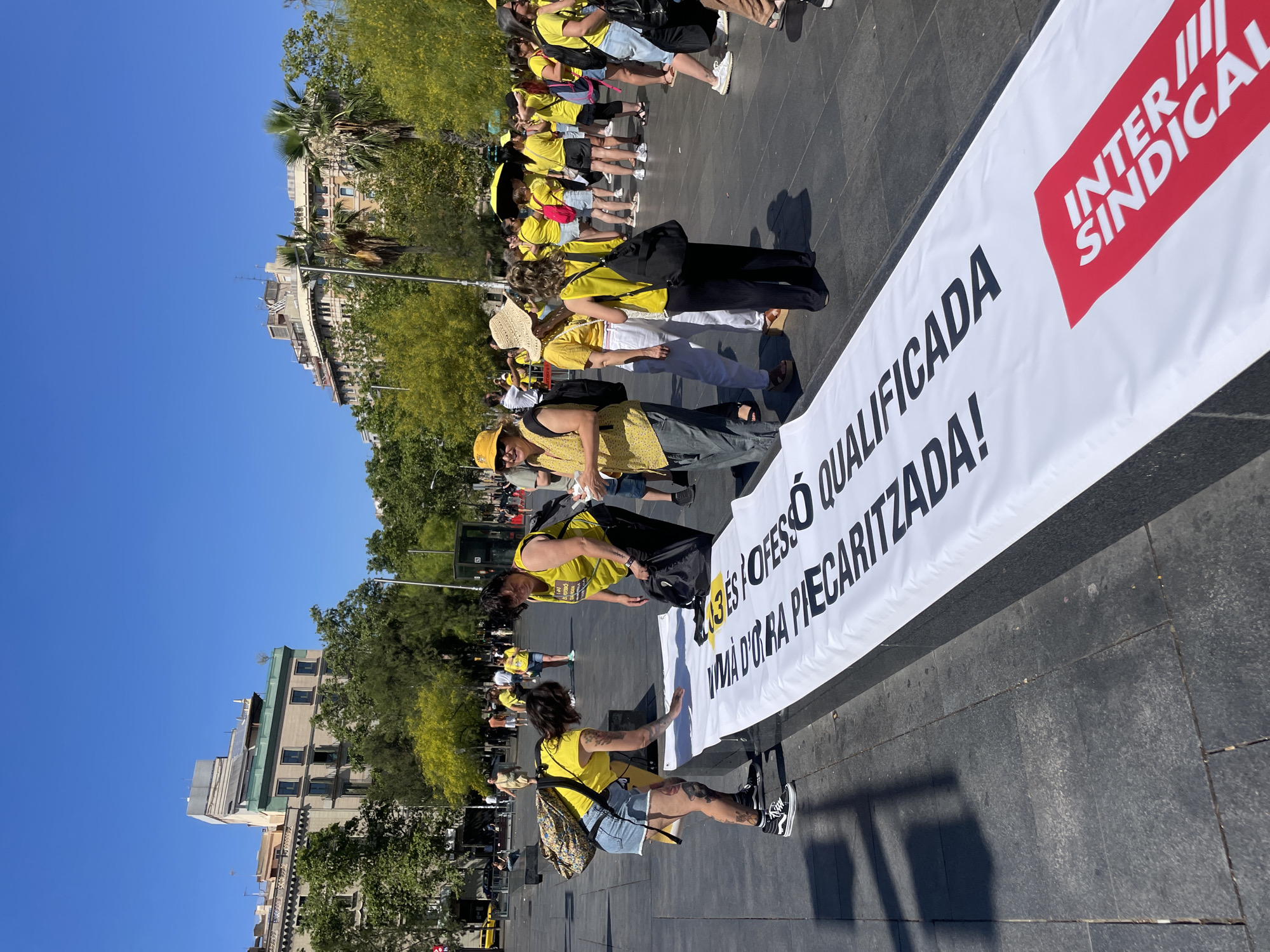 This is a medium-shot, eye-level, outdoor photograph taken on a sunny day, likely in Barcelona, Spain, given the architecture and the Spanish text on the banner. The scene depicts a group of people, predominantly women, gathered around a large white banner laid out on a paved plaza. Many of the individuals are wearing yellow tops and some are wearing hats.
In the foreground, on the left, a woman with tattoos on her legs and arms is wearing a yellow t-shirt, denim shorts, and black and white Vans sneakers. She is looking down and appears to be touching or adjusting the banner. Next to her, a woman in a yellow vest and black pants is also interacting with the banner.
The banner itself is predominantly white with large, bold black lettering and a red logo at the bottom right corner. The text visible on the banner is in Spanish and includes phrases like "INTER SINDICAL" and "QUALIFICADA". The banner is unfurled and extends across the paved ground, which is made of dark grey square tiles.
In the background, more people are visible, some also wearing yellow. The environment includes buildings with balconies, trees with green foliage, and streetlights. The sky is clear and blue. The overall impression is of a gathering or demonstration.