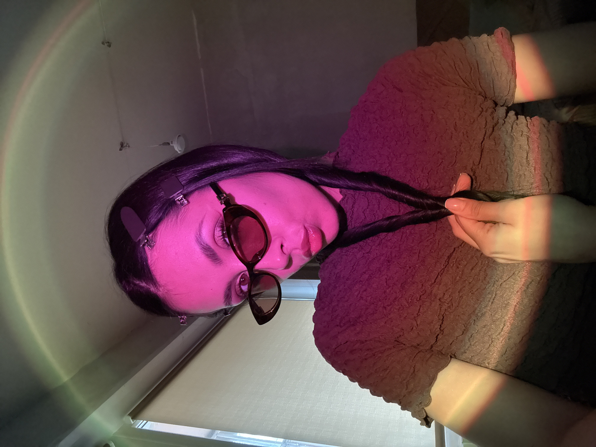 A woman with dark hair is wearing sunglasses and holding two strands of her hair in her hand. She is wearing a textured, reddish-brown top. Her skin is illuminated with a pinkish-purple hue, possibly from a colored light source. The background is partially obscured, but a window with blinds can be seen, suggesting an indoor setting. The lighting casts a rainbow-like effect on the wall in the background. There is no information to suggest the photo was taken in Votkinsk, Russia.