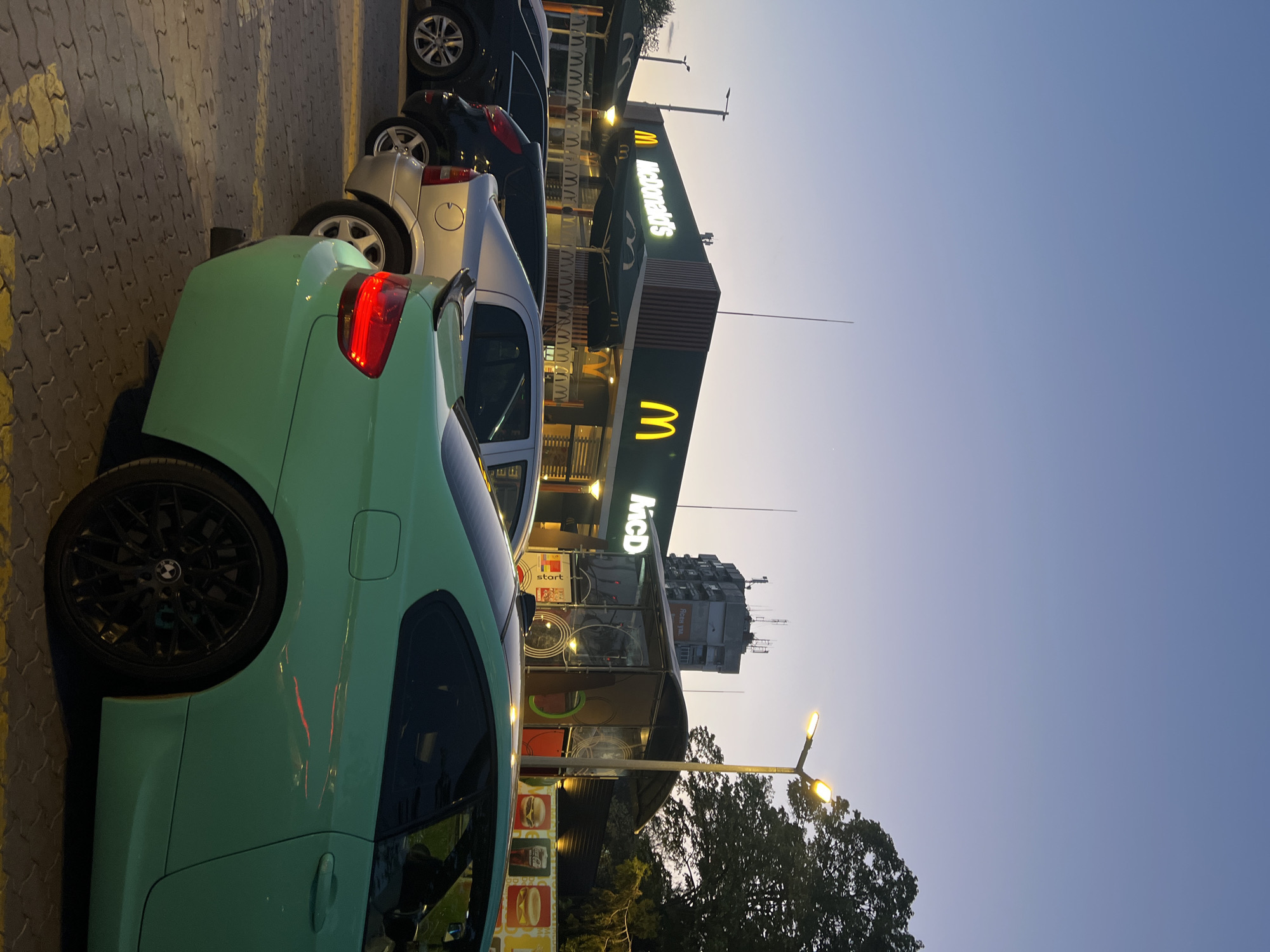 The image depicts an evening scene at a McDonald's restaurant in Plovdiv, Bulgaria, likely a parking lot or drive-thru area. The time of day is dusk or early evening, evidenced by the light blue sky gradually darkening towards the top, and the illumination of artificial lights.
In the foreground, a prominent mint green car is parked on a gray, patterned paved surface with yellow markings. The car's left rear wheel, a black multi-spoke rim with a visible BMW logo, and its bright red illuminated taillight are clearly visible. A small black spoiler adorns the trunk lid. Behind this green car, a silver sedan and other dark-colored vehicles are parked in a line.
The main subject in the mid-ground is the McDonald's restaurant building, characterized by its dark green exterior cladding and multiple brightly lit signs. The iconic "McDonald's" text and large golden arches logos ("M" and "W") are prominently displayed. The building features large windows and what appear to be menu displays or promotional images, including stylized hamburgers, visible through the windows. Umbrellas are also visible near the restaurant, suggesting an outdoor seating area.
In the background, behind the McDonald's building, a tall, older-looking concrete building with several antennas on its roof stands against the fading light of the sky. To the far right, the dark silhouette of trees is visible, partially illuminated by a bright street lamp with two lights that casts a warm glow upwards. The sky is clear, showing no clouds. No people are visible in the scene.