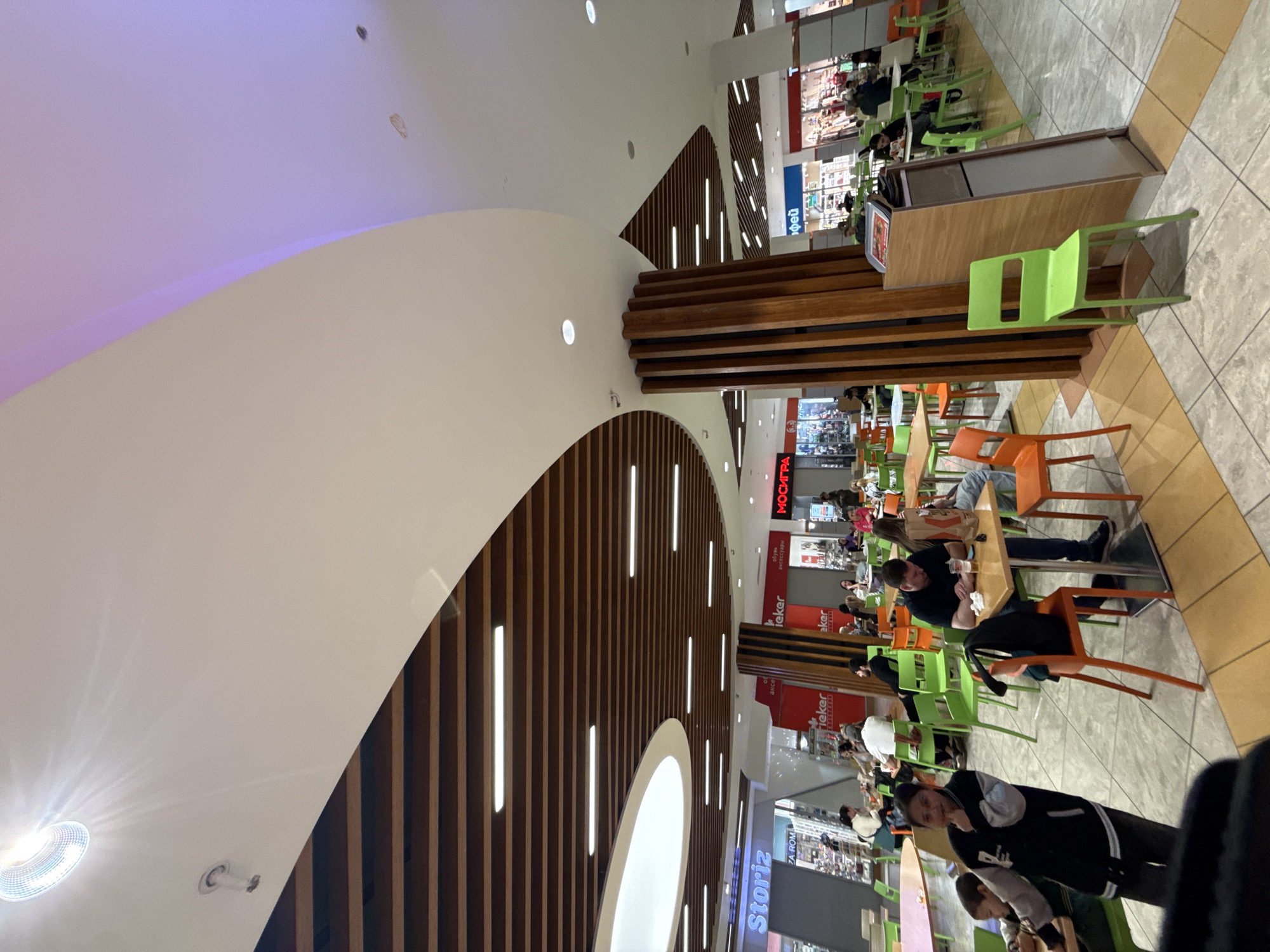 The image shows an indoor food court, likely in a shopping mall, with multiple eateries visible along the upper edges of the frame. Tables and chairs, some occupied, are arranged throughout the area. The architecture includes a curved ceiling with embedded lighting and dark brown linear design elements. People are seen seated at tables, some with food or drinks. The location is likely Moscow, Russia, based on signs written in the Cyrillic alphabet.
