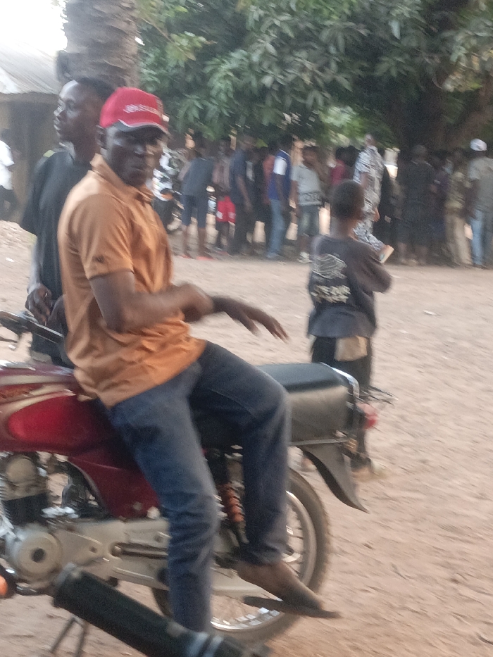 This image captures an outdoor scene in Akwanga, Nigeria, on a reddish-brown dirt surface. In the foreground, a male individual is seated on a stationary red motorcycle with a black seat. He wears an orange short-sleeved polo shirt, blue jeans, black flip-flops, and a red baseball cap with white lettering on the front. His hands are resting on his lap, and his head is turned towards the camera.
Directly behind the primary subject, another male individual in a dark short-sleeved shirt is partially visible. Further into the mid-ground, a child wearing a dark shirt stands with their back mostly to the camera, appearing to hold a white object in their hands. The background shows a gathering of approximately 20 to 30 individuals of various ages and attire, positioned under the canopy of a large tree with green foliage. A prominent, thick, dark tree trunk stands behind the main subject. A rudimentary, light-colored structure is partially visible on the far left edge of the frame. The scene is illuminated by natural daylight.