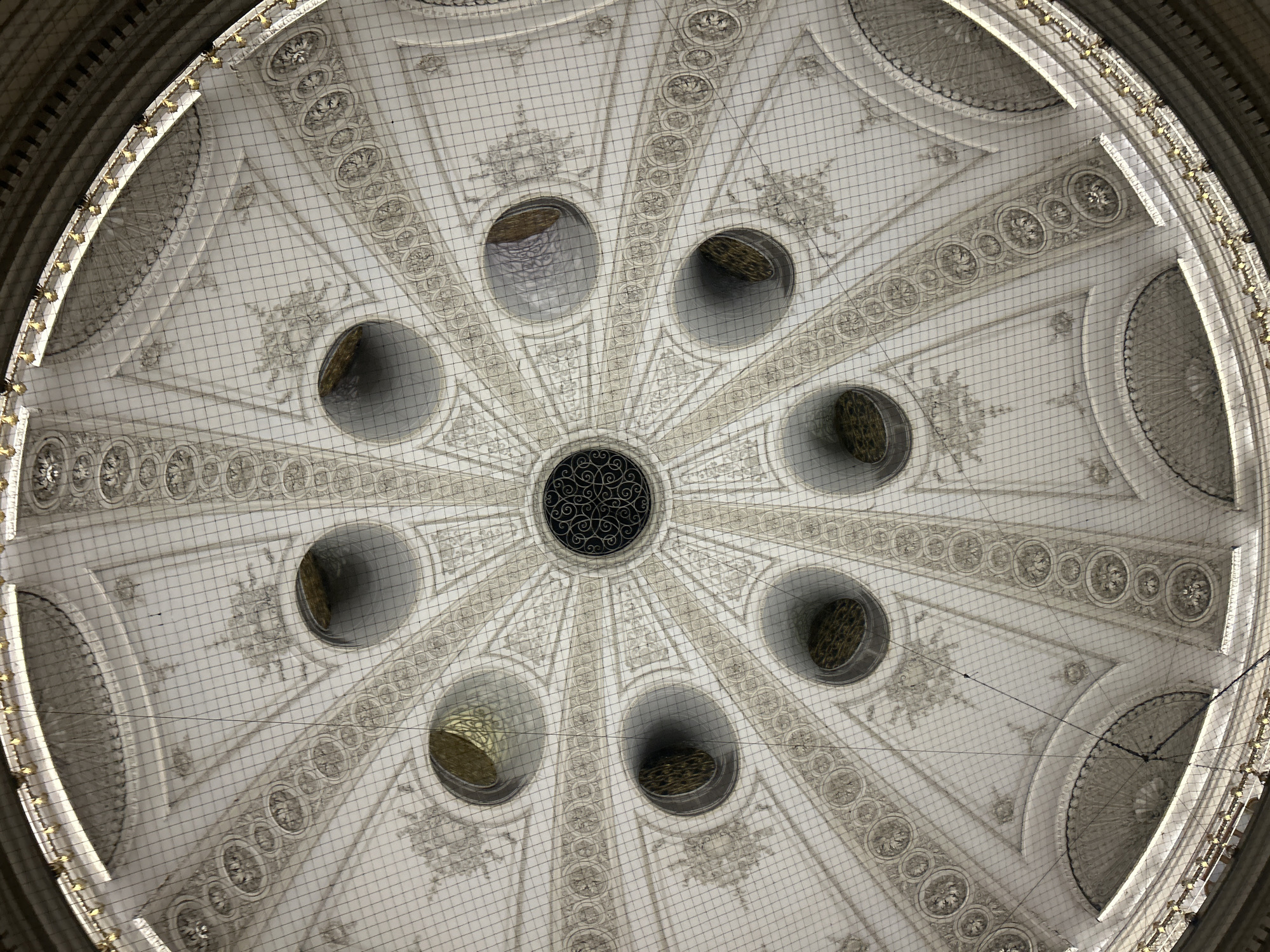 The image shows an interior view looking directly up at the ornate dome of the Austrian National Library (Österreichische Nationalbibliothek) in Vienna, Austria. The dome features intricate white plasterwork with radiating patterns, decorative friezes, and a central circular opening with a decorative metal grille. Surrounding the central opening are eight smaller circular apertures, through which light and gilded decorative elements are visible. The entire dome is covered by a fine mesh netting, likely for safety or protection. The surrounding architecture includes curved sections and a golden decorative railing at the top edge of the visible dome structure. There are no people or visible actions occurring in the scene.