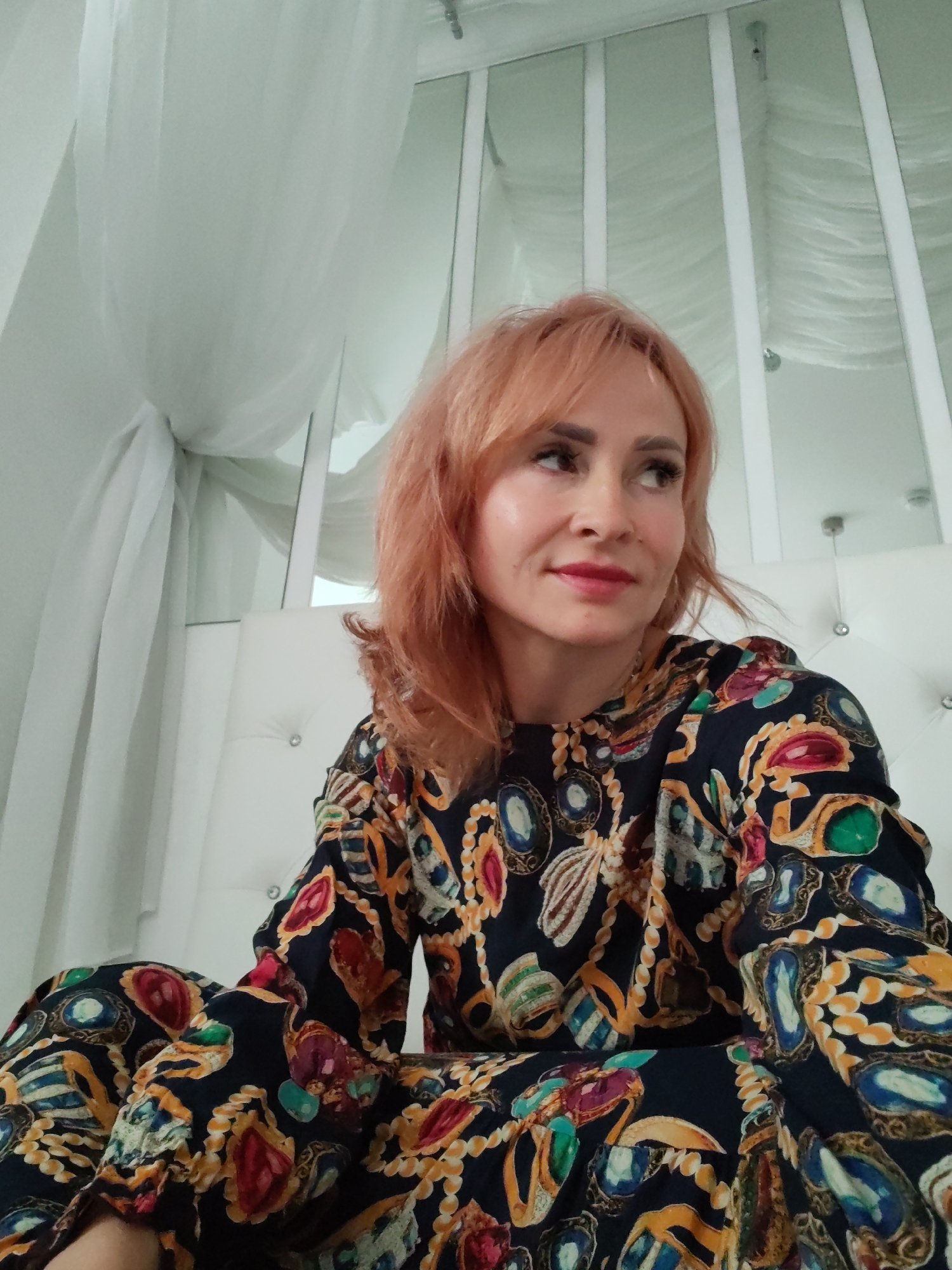This media file depicts a medium shot of a woman with shoulder-length, reddish-blonde hair. She is looking to her left, with a slight smile and her lips slightly parted. She is wearing a dark, long-sleeved dress with a colorful, ornate pattern featuring jewel-like shapes and chains. The woman is seated on a white tufted surface, likely a bed or sofa, which is adorned with crystal-like accents.
Behind her, white sheer curtains are draped, creating a soft, flowing backdrop. Tall, thin white vertical elements with what appear to be mirrors or reflective surfaces are visible between the curtains. These elements suggest a room with a decorative or possibly luxurious interior design. The lighting appears to be soft and diffused, hinting at an indoor setting during the daytime. There are no visible text elements in the image. The woman's gaze and subtle smile convey a relaxed and pleasant emotion. The overall scene evokes a sense of comfort and perhaps an intimate or personal moment.