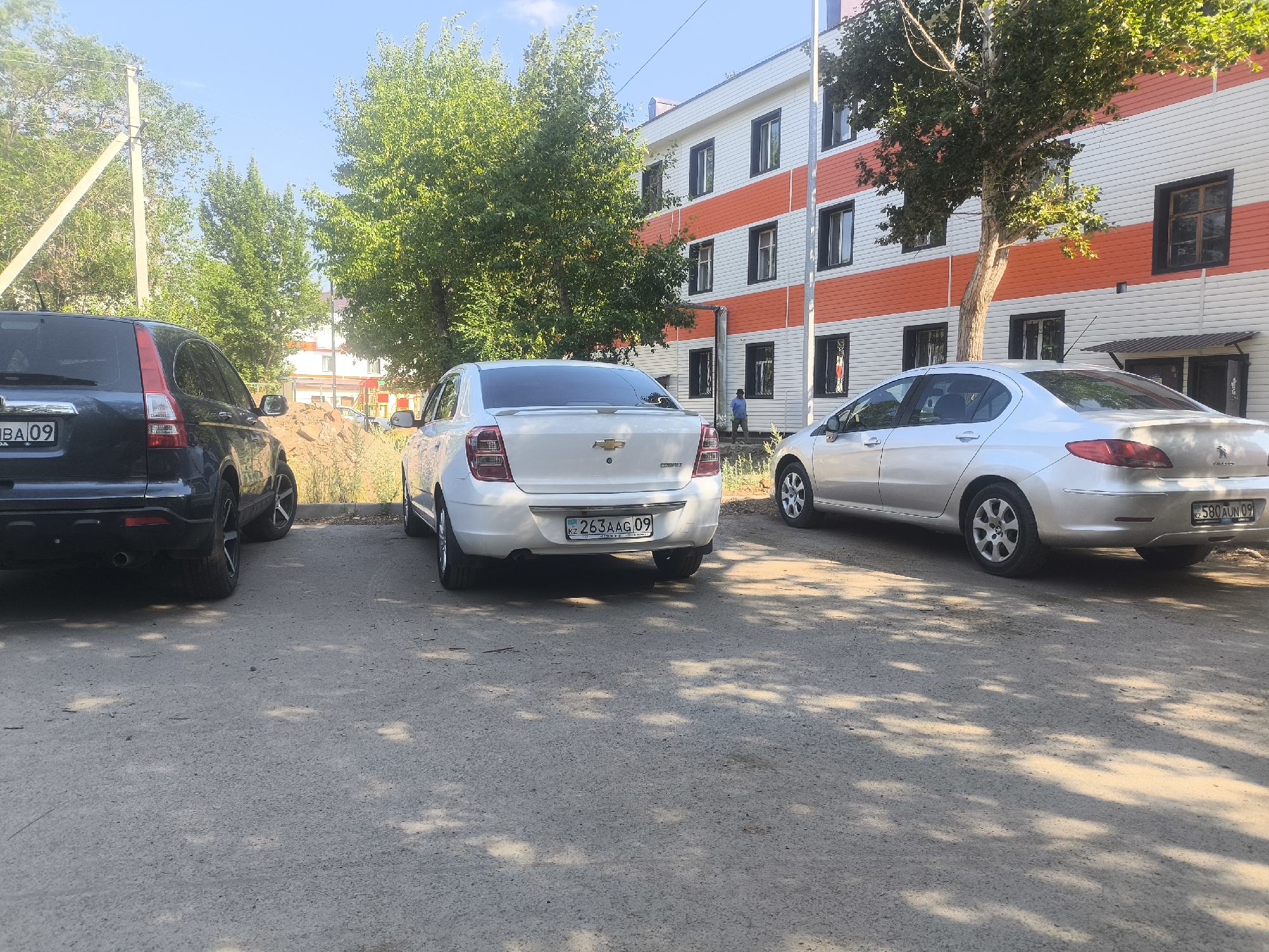 In Abay, Kazakhstan, on a sunny day, a dark-colored SUV is parked facing left on a paved surface. To its right, a white Chevrolet Cobalt sedan is parked, also facing left, with its rear license plate visible, reading "KZ 263AAG 09". Further to the right, a silver sedan is parked, facing left. Behind the vehicles, a building with white and orange horizontal stripes is visible, with several dark-framed windows. Trees with green foliage are present on the left and in front of the building. The ground is a textured asphalt surface dappled with sunlight and shadows from overhead trees. A utility pole with wires is visible on the far left. A person in a light blue shirt is standing near the building entrance on the right.