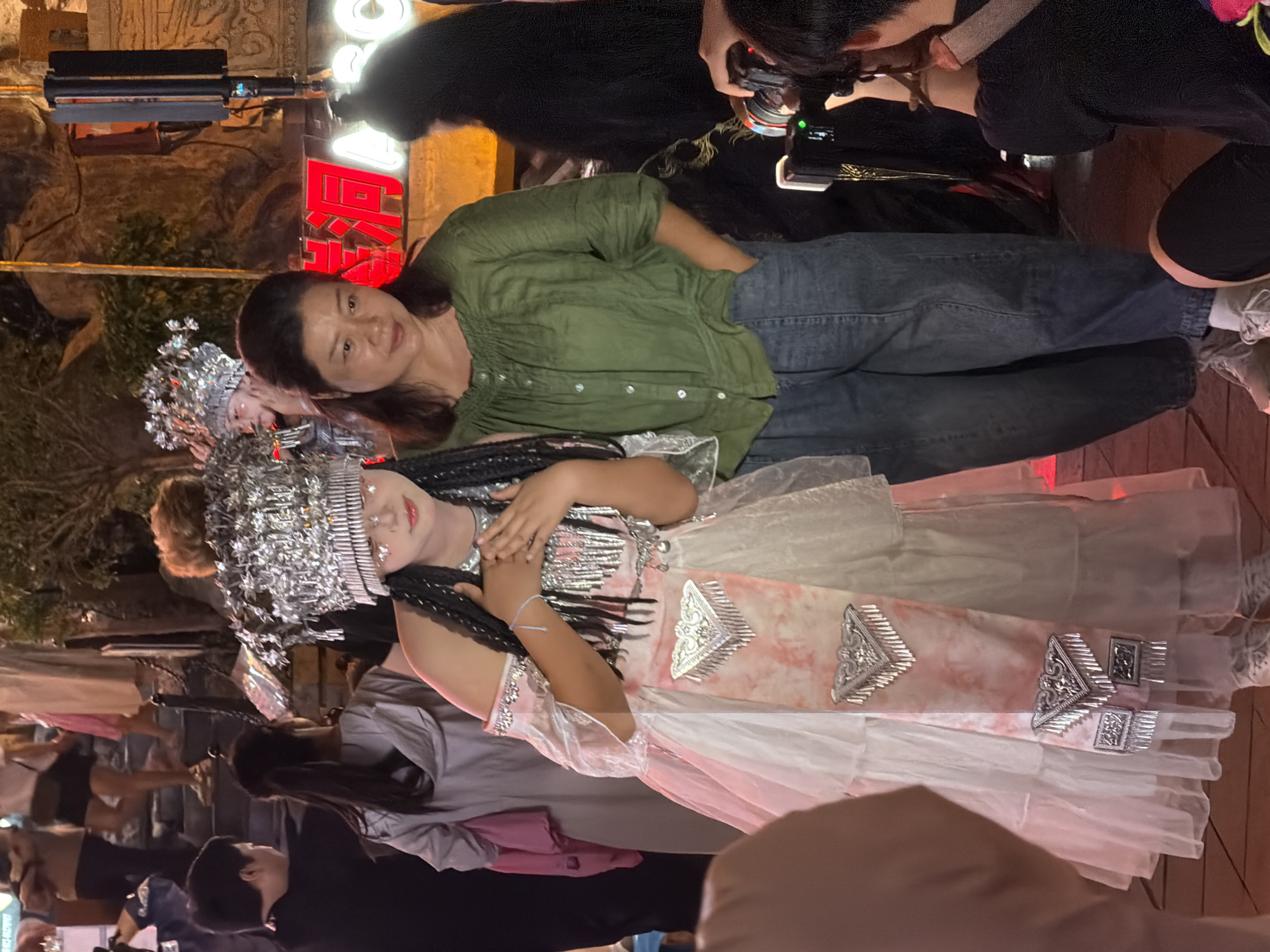 In the Yuzhong District of China, two women are positioned in the foreground, slightly off-center. The woman on the left, wearing a traditional ornate silver headdress and a pink and white tiered dress adorned with silver embellishments, has her hands clasped to her chest. She has braided dark hair and appears to be posing. The woman on the right, wearing a green ruched-neck shirt and blue denim pants, stands with her left hand in her pocket, looking towards the viewer. Behind them, other individuals are visible, some in casual attire, and a person with a camera is actively taking a photo. The background suggests an outdoor setting with ambient lighting and what appears to be a sign with Chinese characters.