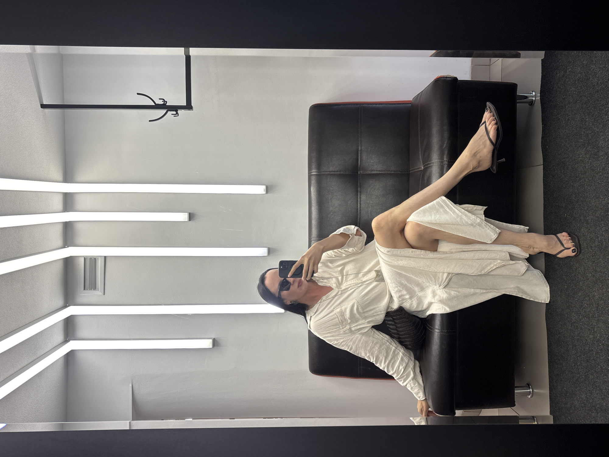 A full-length portrait captures a woman in a reclined pose on a dark leather sofa. She is wearing a long-sleeved white shirt and black high heel sandals. Sunglasses conceal her eyes as she holds a smartphone, apparently taking a selfie. The room's decor is modern, with minimalist lighting fixtures on the walls and a dark floor. A clothes rack is visible in the top left corner. Based on the metadata provided, the location is Chaikovsky, Russia.