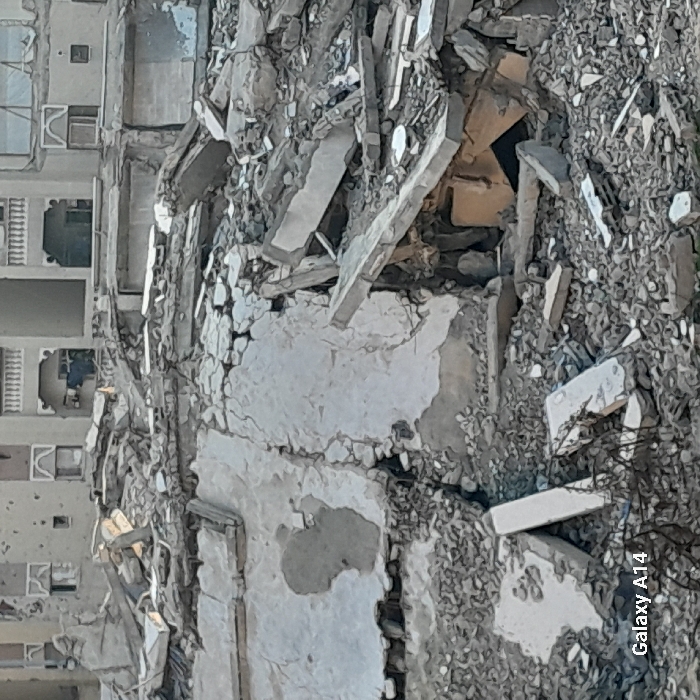 Debris and rubble are piled high, revealing remnants of a destroyed building. Concrete slabs, shattered bricks, and loose gravel dominate the scene. In the background, parts of an adjacent, seemingly undamaged building are visible, showcasing windows and balconies. The destruction contrasts starkly with the standing structure, indicating a catastrophic event. A "Galaxy A14" watermark appears in the corner.
