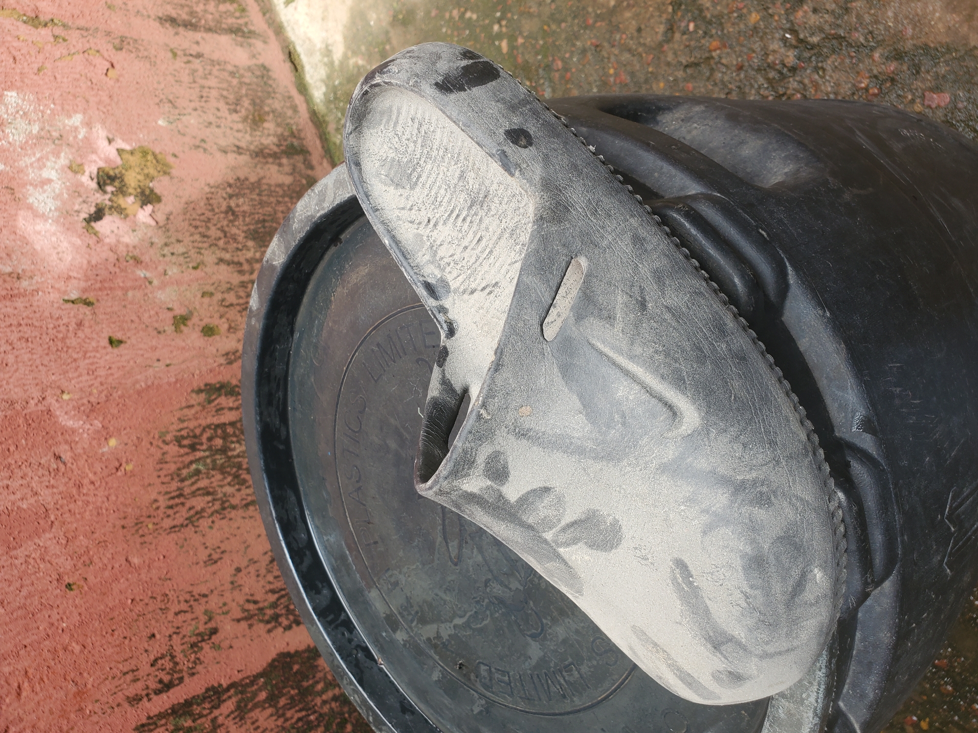 A close-up, overhead shot shows a black plastic sandal resting on a black plastic bin lid. The sandal is dusty, with visible tread marks and scuff marks on its surface. The bin lid also appears dusty and has some moisture or dirt stains. The lid has embossed text on it, but it is difficult to read clearly due to the dust and angle. Partially visible words appear to be "PLASTICS LIMITED." The background consists of a reddish-brown, rough surface, possibly concrete or pavement, with some patches of green moss or algae. The lighting suggests it is daytime, with a slightly overcast or diffused light, as there are no harsh shadows. The overall scene is somewhat unkempt, suggesting an outdoor setting that is exposed to the elements and possibly in use for storage or disposal. The location context is Jos, Nigeria.