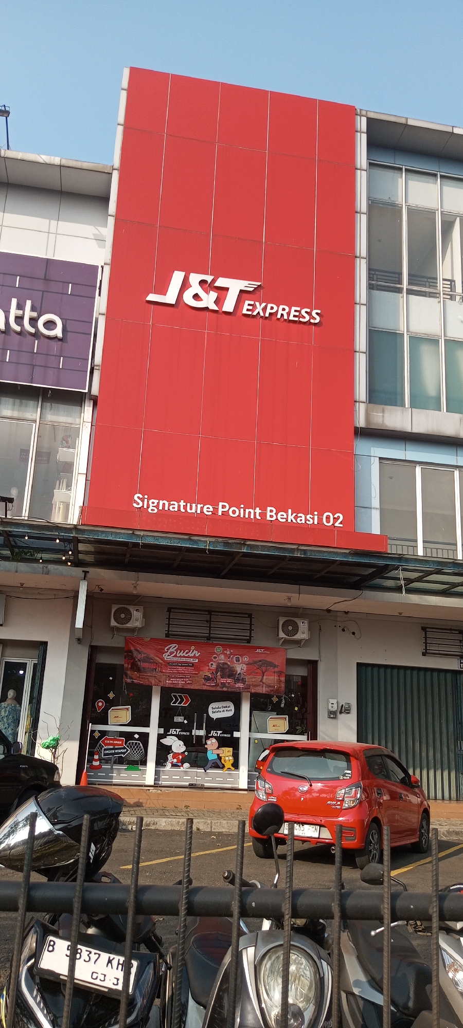 The image captures the exterior of a J&T Express courier service branch, identified by prominent signage. The building features a large red facade with the J&T Express logo displayed prominently. Below the logo, text reads "Signature Point Bekasi 02".
In front of the building, there is a storefront with glass windows displaying graphics and advertisements. One banner reads "Bucin" and features images related to deliveries. Another sign displays cartoon characters and the text "J&T Express". Two air conditioning units are mounted on the exterior wall above the storefront.
A red hatchback car is parked in front of the building, and several motorcycles are visible in the foreground, some behind a metal fence. The environment suggests an urban or commercial area in Lambangjaya, Indonesia, with a clear sky overhead. The overall scene is that of a functional business establishment on a bright day.
