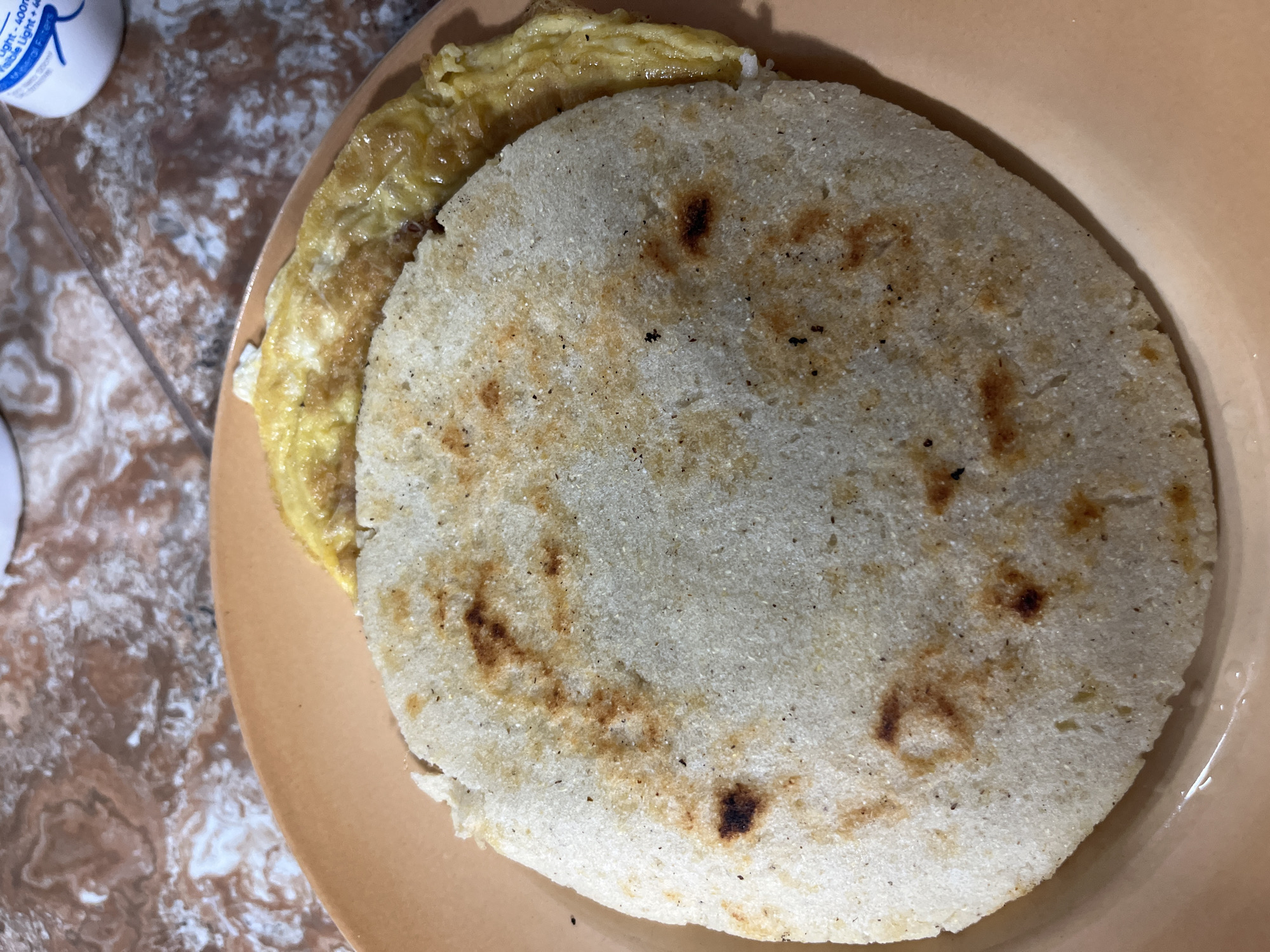 A close-up, overhead shot shows a plate of food on a tiled surface. The plate is a light orange color. On the plate are two food items: a large, circular white corn patty, slightly browned in spots, and a folded omelet, light yellow with some darker brown edges. The tiled surface beneath the plate has a marbled pattern of brown, pink, and white. A white bottle with blue lettering is partially visible in the upper right corner. The image appears to be taken in Juan Griego, Venezuela.
