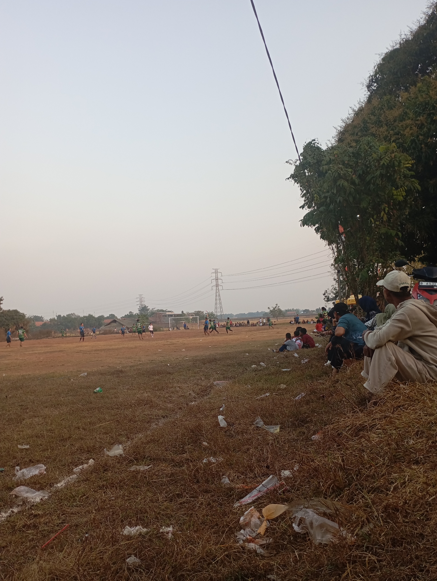 The image depicts a soccer game in progress on a dry, dirt field in Kecamatan Haurgeulis, Indonesia. Several players, distinguished by their blue and green jerseys, are visible on the field, actively participating in the match. A goal net is positioned at the far end of the field. Along the right side of the field, a group of spectators are seated on the ground, some under the shade of trees. One spectator in the foreground is wearing a light-colored hooded sweatshirt and a white baseball cap. The environment is characterized by dry grass, scattered debris including plastic bags and wrappers, and utility poles with power lines in the background. A motorbike is parked near the spectators. The sky is hazy, suggesting it may be late afternoon or evening.