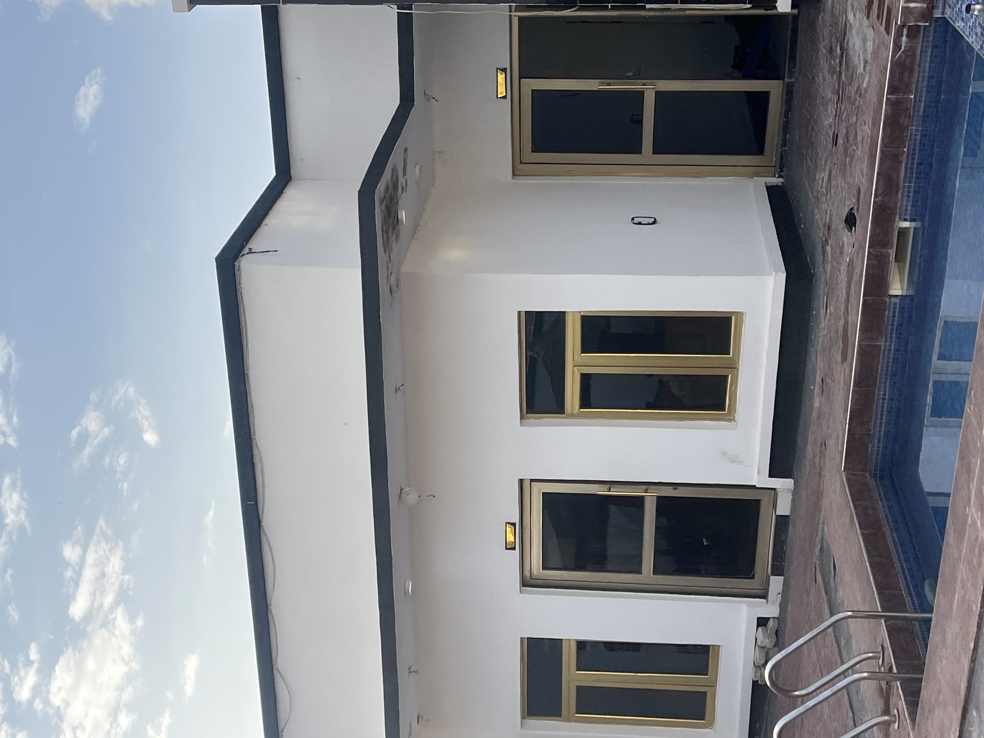 The image depicts the exterior of a modern building in Jauro Yinu, Nigeria, with a small swimming pool in the foreground. The building is painted white with black trim along the roofline and base. It features three large windows with gold frames and dark tinted glass, and two doors, also with gold frames and glass panels. One door is a single panel, while the other appears to be a double door or has a large glass panel on the side. Above one of the doors, a rectangular sign with a yellow light is visible. A security camera is mounted on the exterior wall. The swimming pool is tiled with blue mosaic tiles and has a dark red coping. Stainless steel handrails are installed on the side of the pool. The surrounding ground is paved with dark tiles. The sky above is blue with scattered white clouds. There are no people visible in the image.