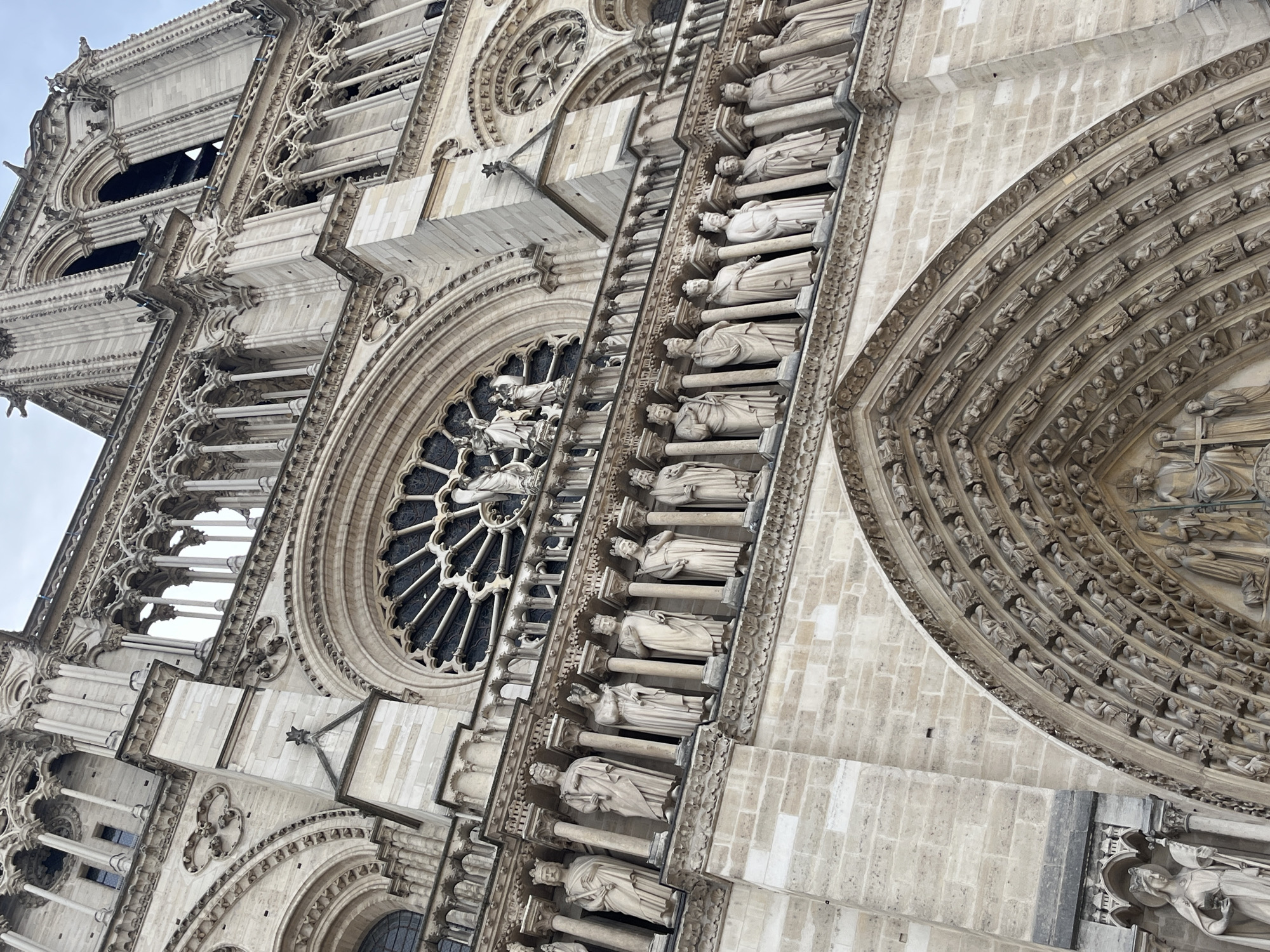 The image shows a low-angle, close-up view of the facade of Notre Dame Cathedral in Paris, France. The cathedral is constructed of light-colored stone and exhibits intricate Gothic architecture. Numerous statues of figures, likely kings or saints, line a prominent balcony, each standing within its own carved niche. Above this balcony, a large, circular rose window with stained glass is visible, surrounded by detailed stonework. Further up, more architectural features, including columns, arches, and spires, extend towards an overcast sky. The overall impression is of grandeur and historical significance, with meticulous craftsmanship evident in every detail.