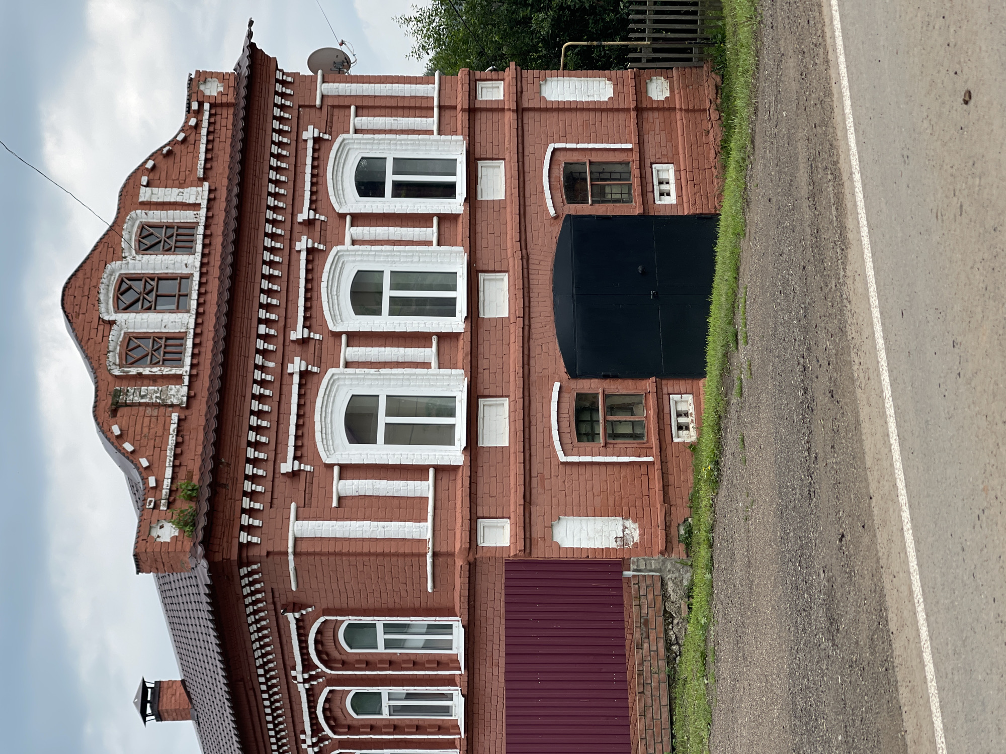 A medium shot captures the exterior of a two-story brick building on a cloudy day. The building is constructed from red brick with white accents around the windows and along the roofline. The second story features several arched windows with white frames, while the first story has a large, dark garage door and a smaller window with a barred pane. A dark-colored metal fence runs along the left side of the building, partially obscuring a section of the first floor. The building sits on a patch of green grass, with a gravel road and a paved road with a white line in the foreground. Trees are visible in the background to the right of the building. The sky is overcast and gray.