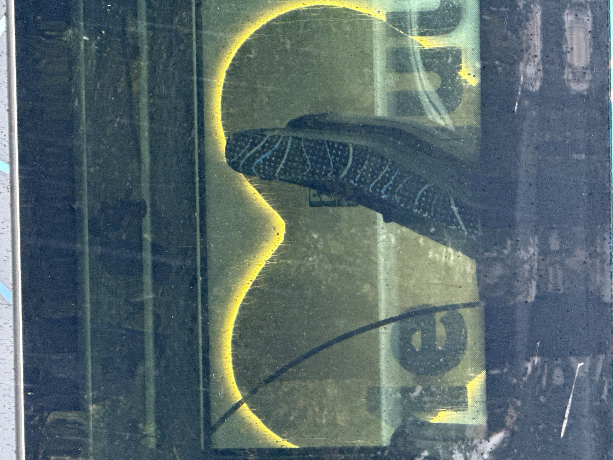 The image shows a close-up view through a dirty glass window, possibly of a vehicle. A shoe with a distinctive blue and white pattern is visible, resting against a illuminated yellow outline of a heart shape on a light-colored background. Parts of text, possibly "te" and "ut", are also visible on the light background. The scene appears to be outdoors, as suggested by the reflections and grime on the glass. There is no specific indication of the city of Tallinn, Estonia.