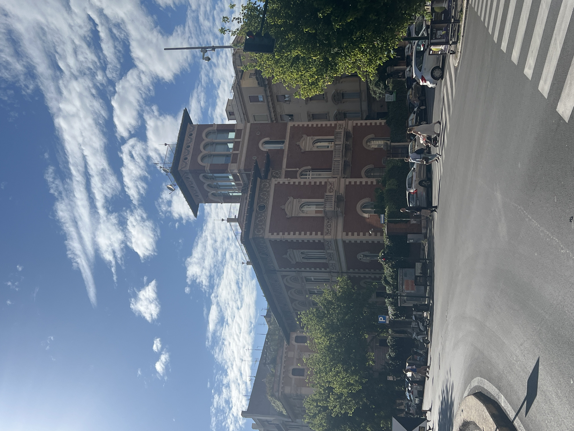 This image depicts a sunny urban street scene at the intersection of Via Saverio Mercadante and Via Po in Rome, Italy. The prominent building dominating the right side of the frame is a multi-story structure with red brick facades and light-colored classical architectural detailing, including arched windows, ornate cornices, and quoins. A distinctive, lighter-colored tower with an open loggia featuring arched windows tops the building at the corner, with satellite dishes and various antennae visible on its roof.
To the left and behind this building, other multi-story residential buildings, mostly light-colored, are visible. Some greenery and small trees grow on the roofs or upper levels of these structures.
The street itself is asphalt, curving from the foreground towards the left and center. A white zebra crossing is visible in the foreground on the right side. Strong sunlight, originating from the upper left, casts distinct shadows on the road, from trees, and from the buildings.
Several individuals are present on the street. On the right, three people are crossing the zebra crossing: a woman in a light dress pushing a baby stroller, a man in a light shirt and dark trousers, and another person partially obscured. Further to the left, a man in a dark shirt and light trousers walks towards the left side of the frame in the street. Another man is walking away from the camera on the far left pavement.
Multiple vehicles are parked along the street on both sides, including cars of various models and colors, and several mopeds or scooters on the sidewalk. Green deciduous trees with full foliage line portions of the street and obscure the lower levels of some buildings. Street furniture includes a black traffic light pole on the right, other utility poles, a blue 'P' parking sign, and a small advertising kiosk on the right pavement. The sky is bright blue with scattered white cumulus and cirrus clouds.