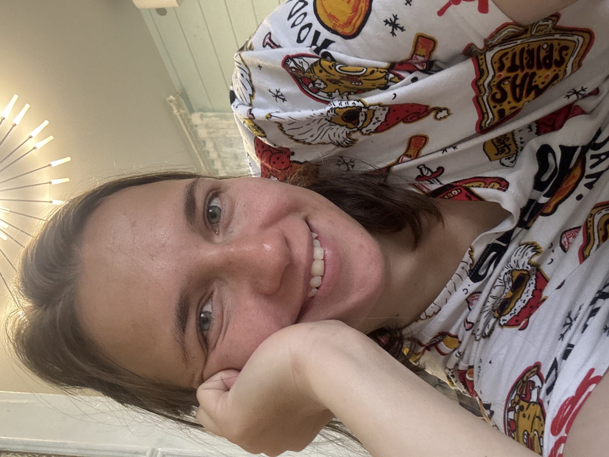 A young woman is smiling at the camera while resting her face on her hand. She appears to be indoors, with a light fixture visible in the upper left corner and a textured wall behind her. She is wearing a white patterned shirt featuring images of Santa Claus and liquor bottles. The location is not discernible based on visual information alone.