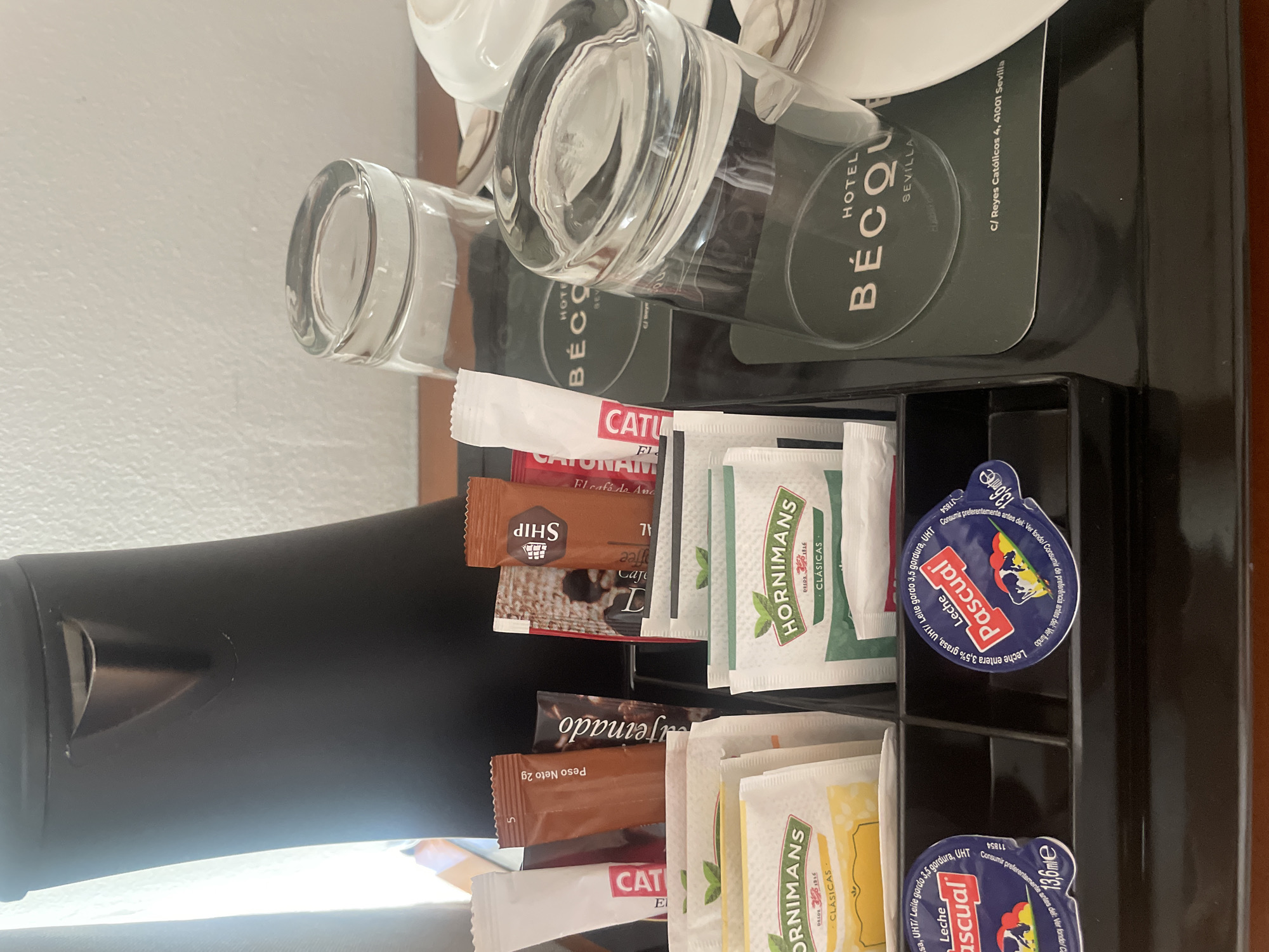 A black electric kettle sits on a dark wooden surface next to a tray of amenities. The tray contains several packets of tea and coffee, two glasses, and two small containers of milk. One of the glasses is upside down on a dark green coaster that reads "HOTEL BÉCQUE SEVILLA C/ Reyes Católicos 4, 41001 Sevilla". The other glass is upright and filled with a clear liquid. The milk containers are blue and white with red lettering and depict a cow. The packets of tea and coffee have various branding, including "HORNIMANS" and "SHIP". The background shows a textured white wall and a light-colored curtain.