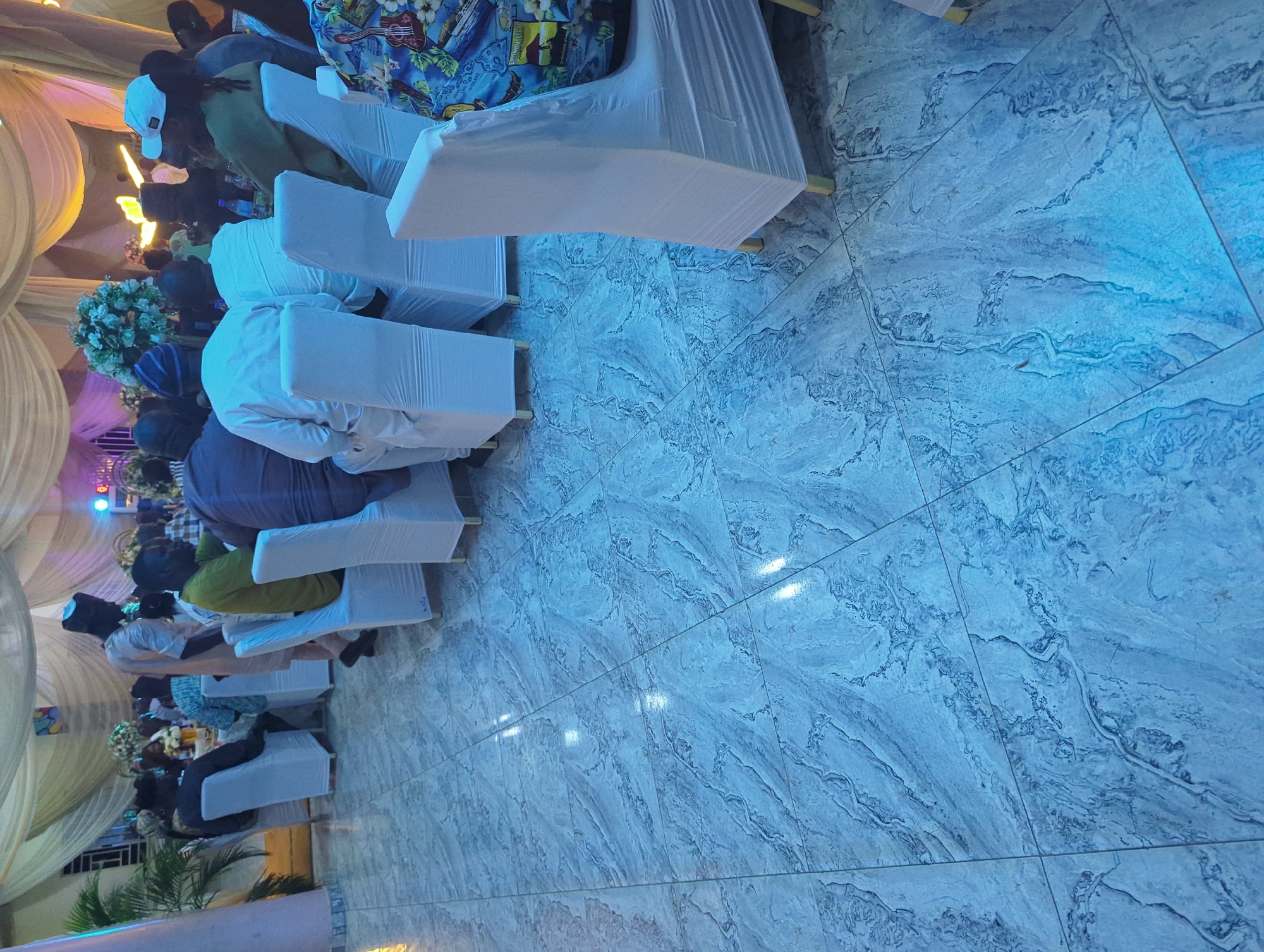The image captures an indoor event, possibly a reception or gathering, with guests seated at tables covered by white cloths. The floor is tiled with a marble-like pattern in shades of blue and gray, reflecting ambient light. In the foreground, several chairs are draped with white slipcovers, and a portion of a person wearing a colorful Hawaiian-style shirt is visible on the right. In the middle ground, men are seated and standing, some wearing traditional African attire. The background features draped curtains and floral arrangements, suggesting a decorated venue. The overall lighting is dim with accents of blue and yellow. The image does not provide any specific landmarks or visual cues to definitively place the location in Barden Yana, Nigeria, although the attire of some individuals might suggest a Nigerian context.