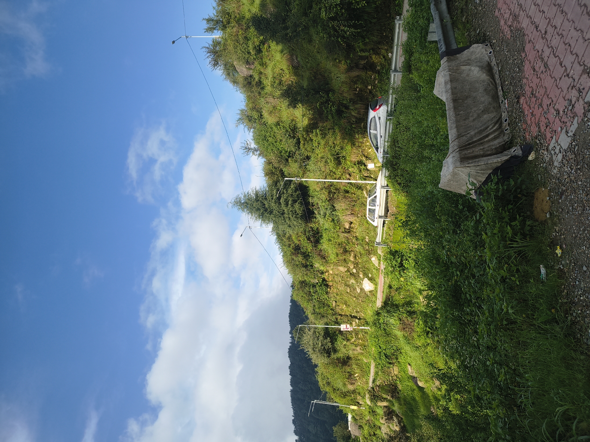 The image depicts a roadside scene in Haini, India, under a bright blue sky with scattered clouds. A winding road curves uphill, bordered by lush green vegetation and rocky outcrops. Two cars, a white sedan and a silver hatchback, are visible on the road. Several streetlights are positioned along the roadside. In the foreground, a tarp covers an object next to a paved pathway and a gravel area. The overall environment suggests a mountainous or hilly region with abundant greenery.