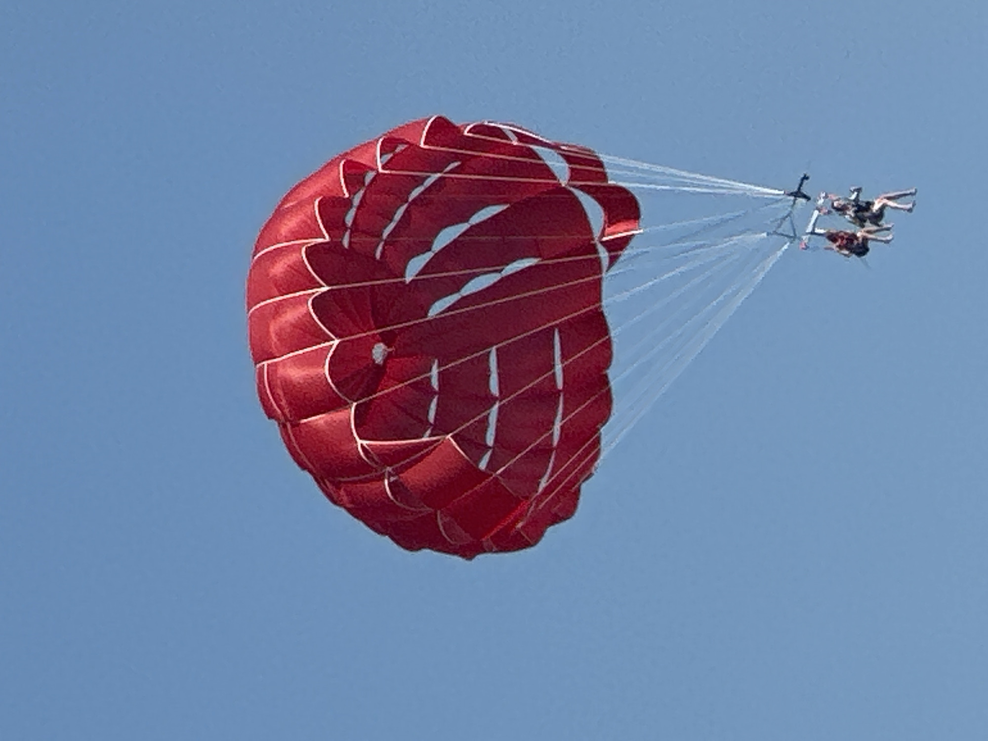 In the sky above Boreti, Montenegro, a red parachute is in flight, carrying two individuals. The parachute, a vibrant red with white accents, is inflated and taut, suspended against a clear blue sky. Attached to the parachute by thin lines is a harness system from which two people are suspended. They appear to be seated, enjoying the aerial experience. The overall scene is one of recreational activity set against a serene natural backdrop.