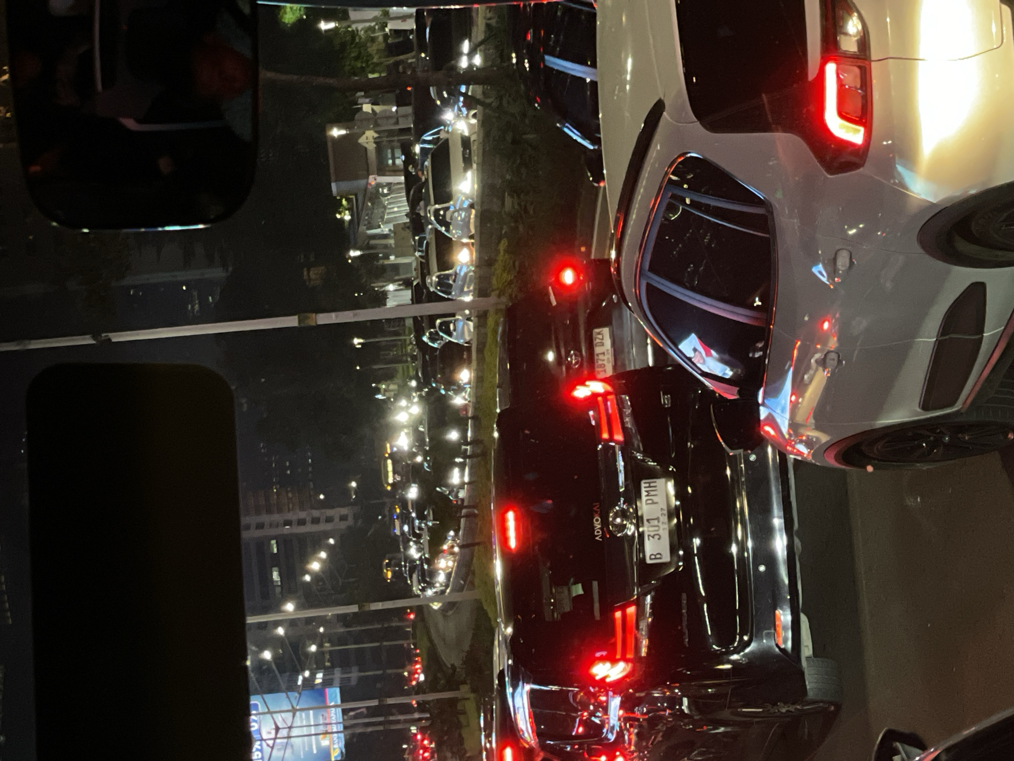 The image, taken at night, depicts a traffic jam on a street in South Jakarta, Indonesia. Cars are lined up bumper to bumper, with their tail lights illuminated. Streetlights and building lights are visible in the background, contributing to the overall brightness of the scene. The vehicles and other objects are also viewed in a car's passenger-side mirror.